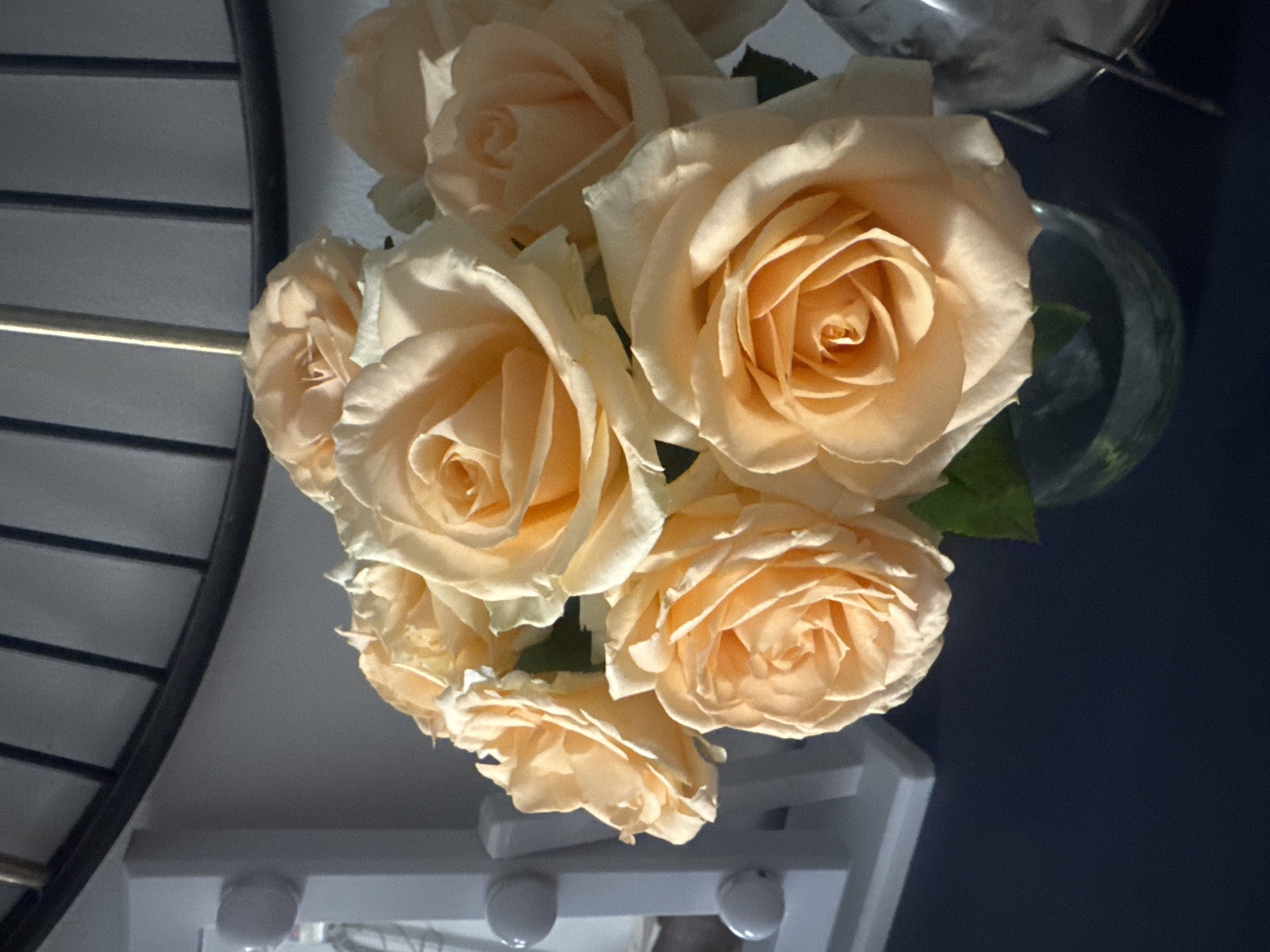 A cluster of light peach-colored roses in a glass vase rests on a white surface. The roses are in various stages of bloom, with some fully open and others tightly budded. A dark blue wall is in the background, and part of a mirror with a black frame is visible on the left. The lower portion of the white surface appears to be a vanity with round knobs. The scene is indoor. There is no evidence to support the image being located in Bat Yam, Israel.