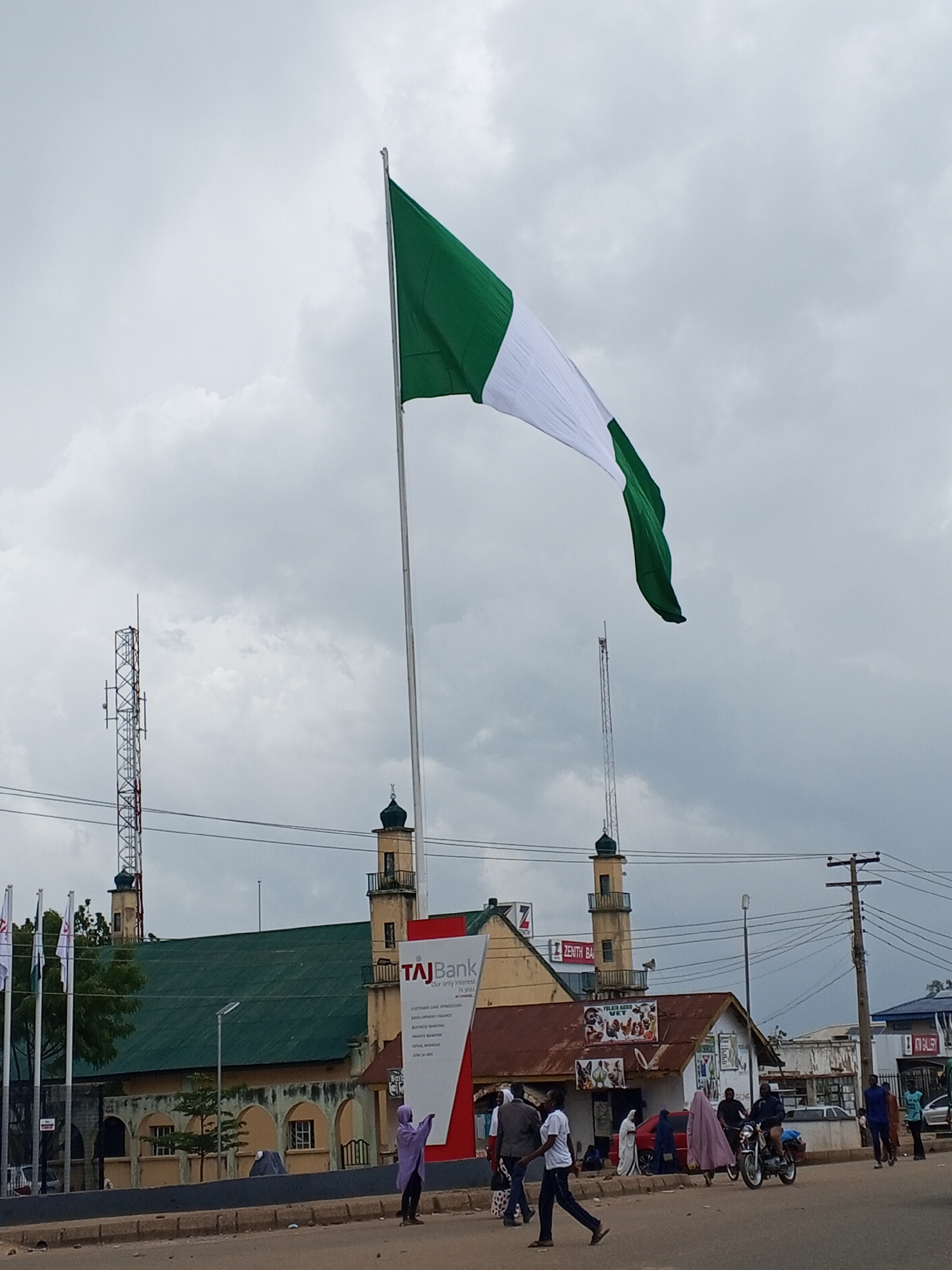 A Nigerian flag flies from a tall flagpole in Bukuru, Nigeria, against a cloudy sky. In the background, a mosque with two minarets is visible. Several flags on poles stand to the left of the mosque. A TAJ Bank sign is positioned in front of the mosque. On the street in front of the buildings, several people are walking and a motorcycle is being ridden by two individuals. A telecommunications tower is also present in the scene.