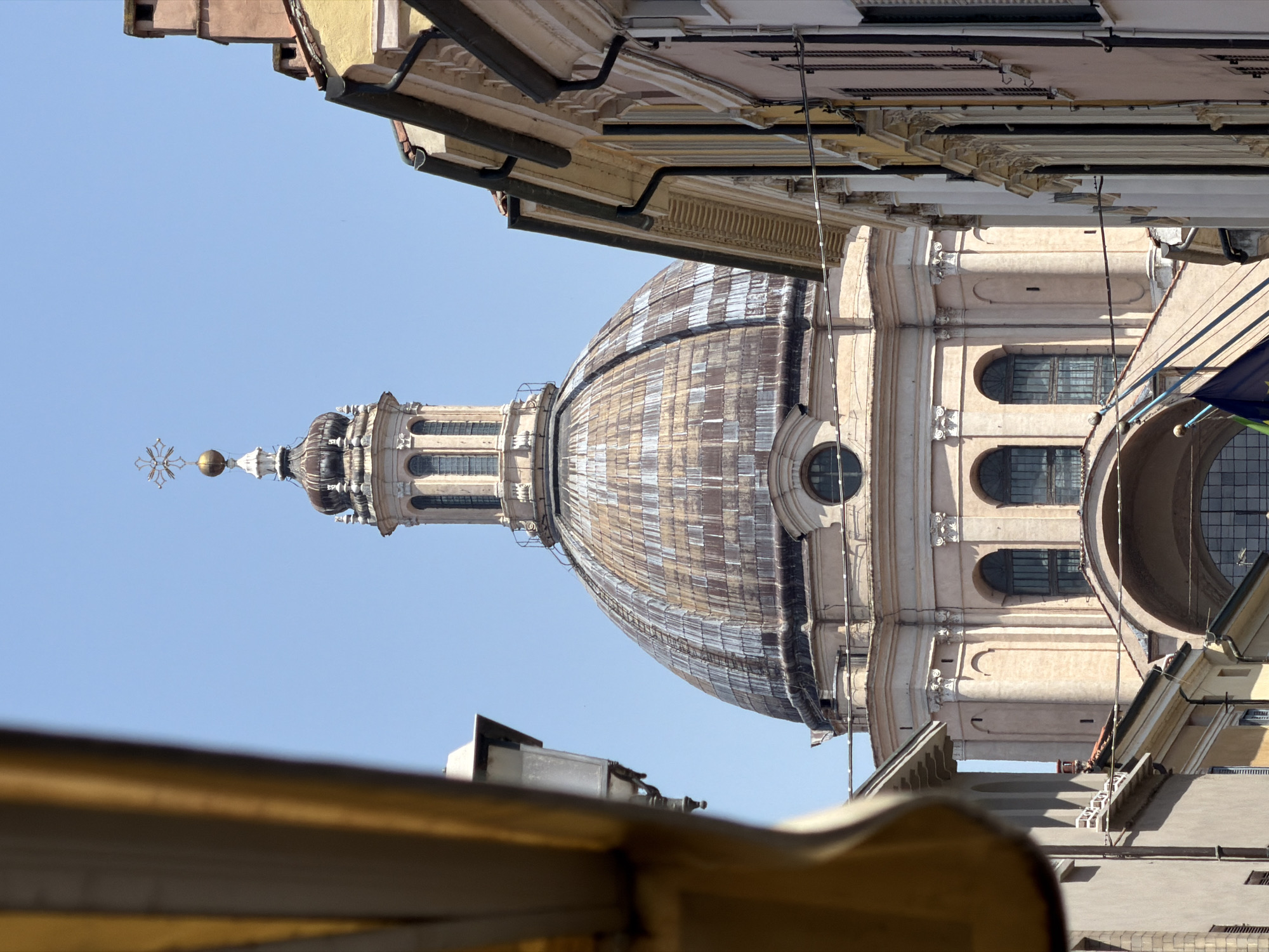 Looking upward, the image focuses on the dome of Basilica di Sant'Andrea from a lower vantage point within the city of Mantua, Italy. The dome, featuring a patterned surface and an ornate lantern, rises against a clear, light blue sky. Surrounding structures with diverse architectural elements, including roofs and facades, frame the dome from above. An awning-like structure appears in the lower foreground.