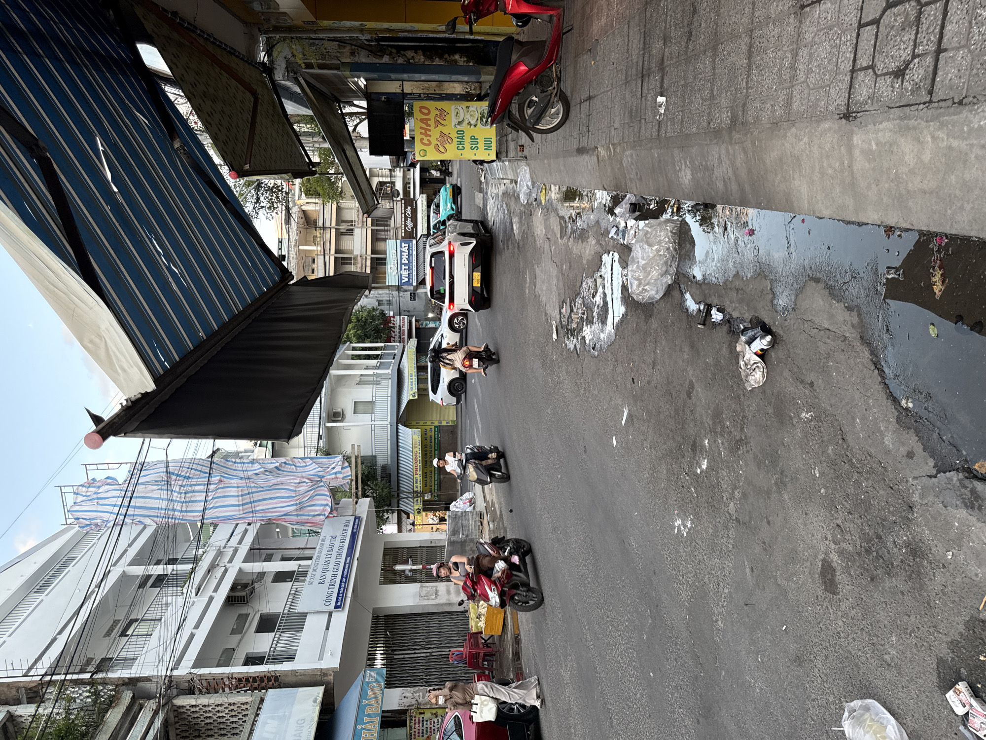 The image depicts a street scene in Nha Trang, Vietnam, characterized by a mix of commercial buildings, residential structures, and vehicular traffic. The road surface is asphalt, showing signs of wear and tear, with visible puddles and scattered debris, including plastic bags and wrappers, suggesting a lack of regular street cleaning.
On the left side of the street, a multi-story white building is under construction, covered partially with blue and white striped scaffolding netting. A sign on the building indicates it is the "DEPARTMENT OF CONSTRUCTION KHANH HOA" and details an ongoing project. Further down the street, a red car is parked. A woman in light-colored clothing is walking away from the car towards the camera, carrying a white bag and wearing a face mask.
The street is active with motorbikes. Two individuals on motorbikes are moving towards the camera: one on a red motorbike, wearing a helmet and a light-colored top, and another on a dark-colored motorbike, also wearing a helmet. In the background, other vehicles, including a white SUV and a teal car, are visible on the road.
Overhead, awnings of varying designs are extended from the buildings, providing shade. One prominent blue and white striped awning is partially obscured by the frame. On the right side of the street, a yellow sign with Vietnamese text, likely advertising food, is displayed. A red motorbike is parked on the sidewalk near the sign. The sidewalk is paved with concrete tiles.
The overall atmosphere suggests a typical urban environment in Vietnam, with a blend of ongoing development, commercial activity, and the presence of street litter. The time of day appears to be late afternoon or early evening, with the sky a pale blue.