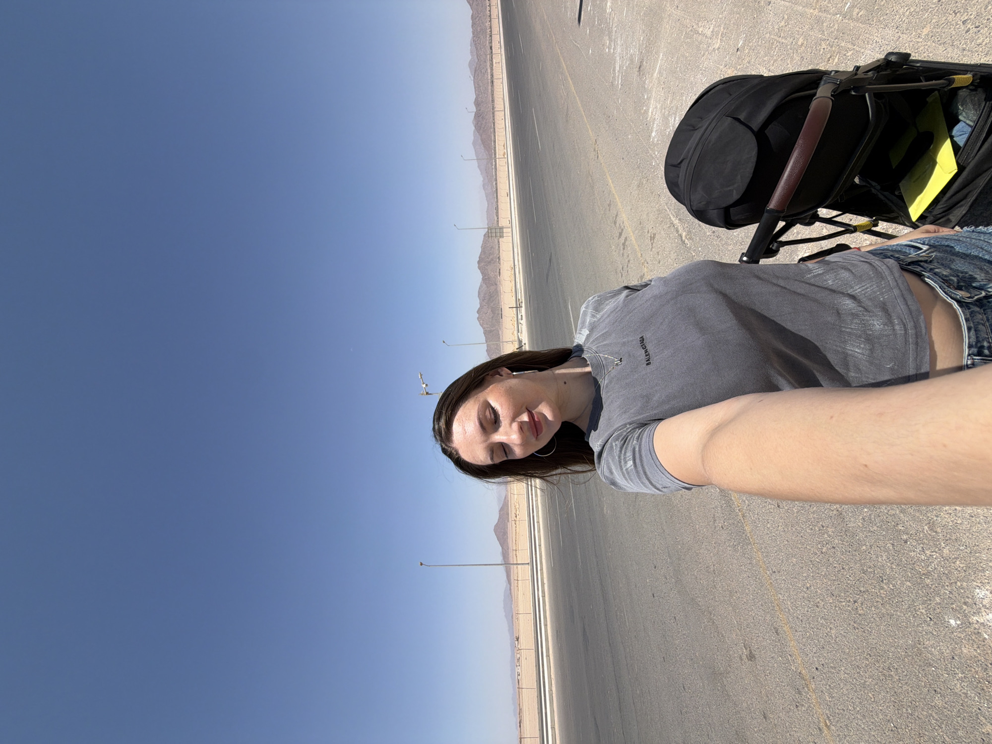 A young woman with long brown hair and closed eyes stands in a selfie pose on a paved road in a desert landscape under a clear blue sky. She is wearing a gray t-shirt and denim shorts. Next to her is a black stroller. In the background, there is a fenced-off area with sand and distant mountains. Streetlights are visible along the road.