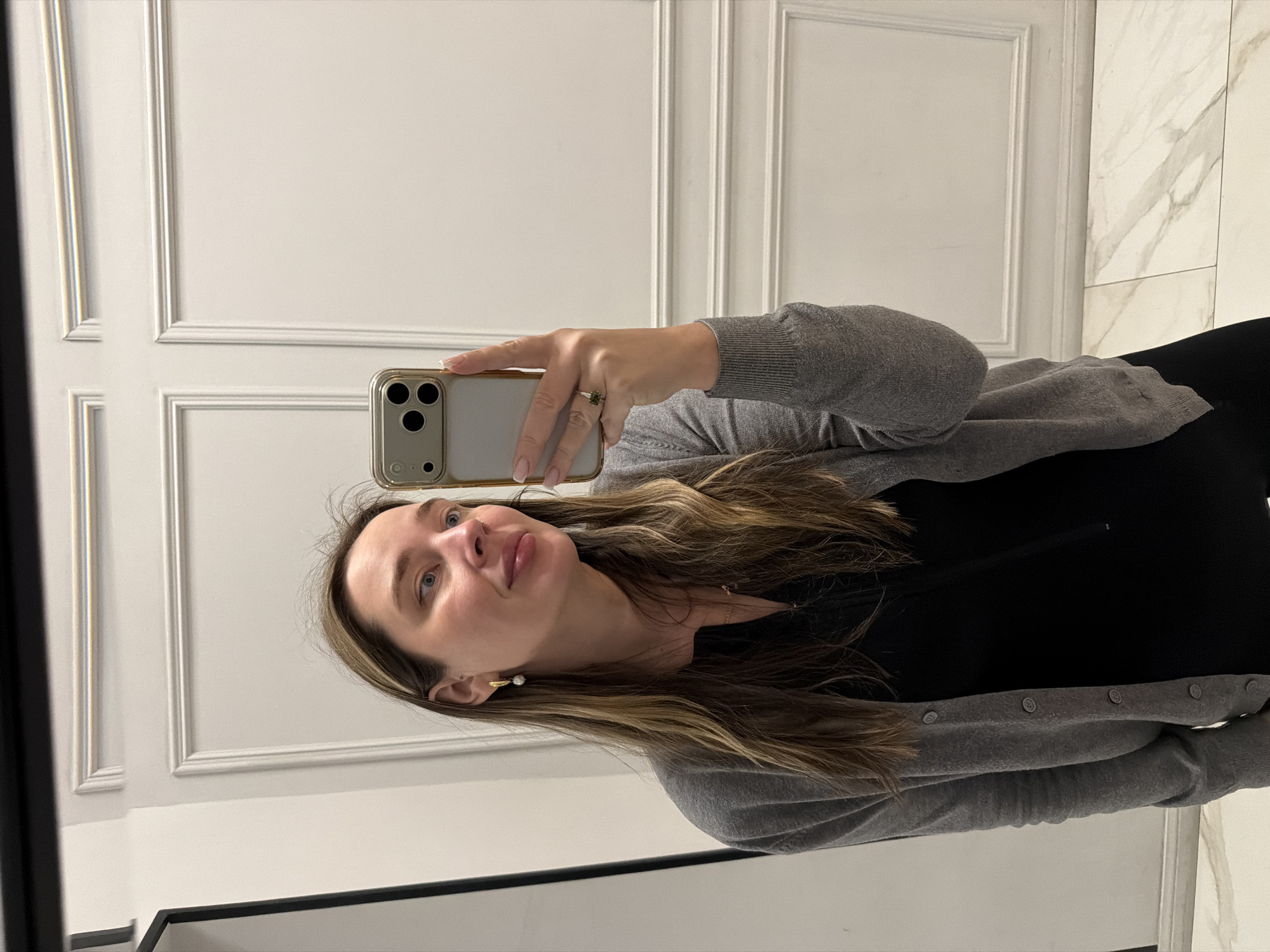 A woman is taking a selfie in front of a wall with paneled and marble sections. She is wearing a black shirt and a gray cardigan. She has long brown hair and is holding a smartphone to take the picture. She appears to be indoors. The location and city information provided are not directly observable in this image.