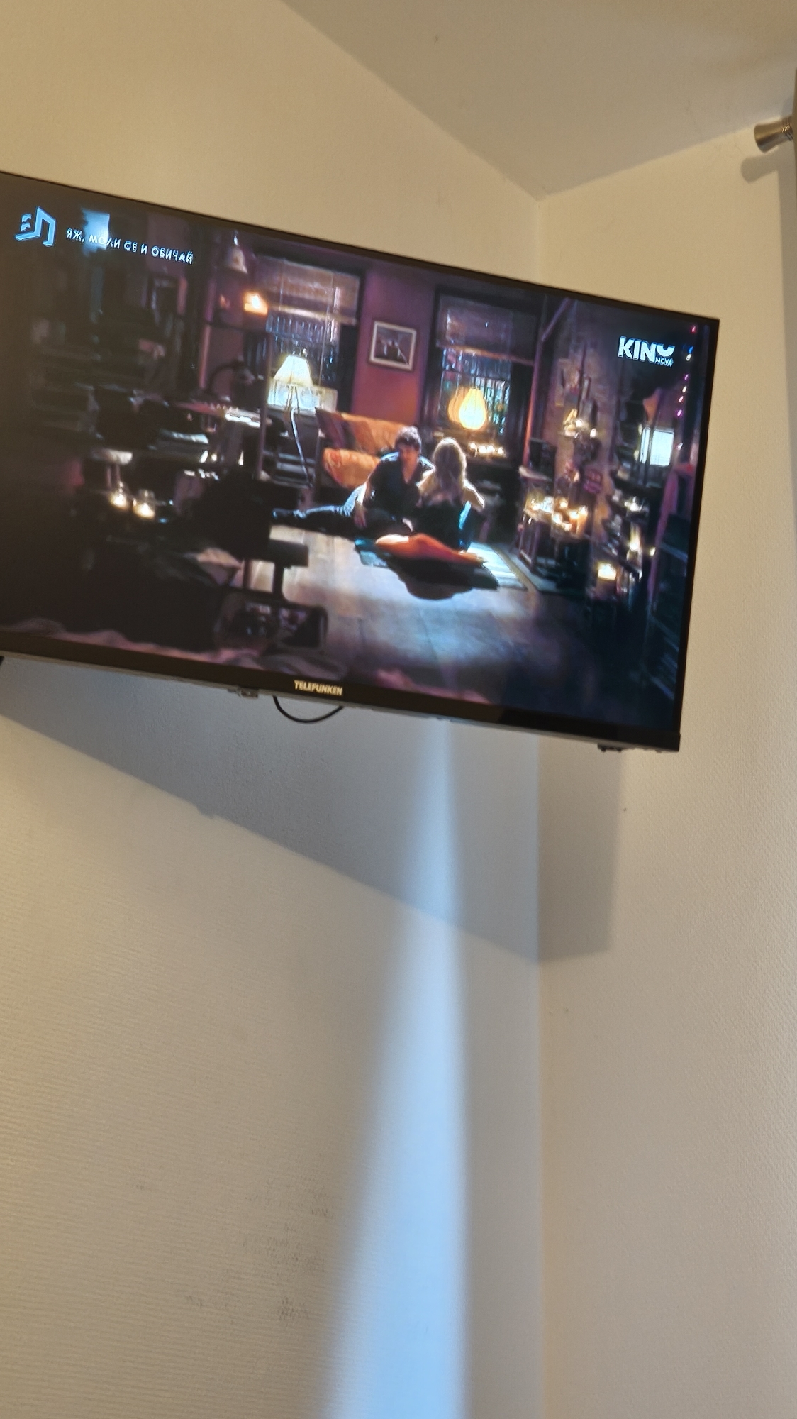 The image captures a television screen displaying a scene from a movie or television show, mounted on a light-colored wall. The main subjects on the TV screen are two individuals, a man and a woman, positioned on the floor indoors. They appear to be interacting, with the man lying down and the woman sitting near him. The setting within the TV scene is an interior room, possibly a living room or study, with warm ambient lighting provided by lamps, and various decorative items or furniture in the background.
Visible text on the television screen includes a Cyrillic phrase in the top left corner, "ЯЖ, МОЛИ СЕ И ОБИЧАЙ," which translates to "EAT, PRAY, AND LOVE," indicating the movie "Eat Pray Love." In the top right corner, the channel logo "KINO NOVA" is visible. At the bottom edge of the TV, the brand name "TELEFUNKEN" is clearly displayed.
The wall on which the television is mounted is plain white, with a subtle textured surface. A faint blue light reflection is visible on the lower part of the wall, suggesting ambient lighting in the room where the photograph was taken. In the upper right corner of the overall image, a metallic fixture or rod is partially visible. The photograph is taken from a slightly upward angle, showing the TV and the upper part of the wall.