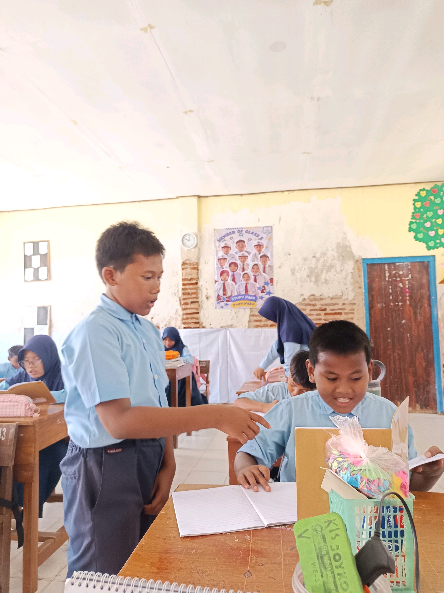 The image depicts a classroom scene, likely in Labuhan, Indonesia, with several students and a teacher. In the foreground, two young boys are the primary focus. One boy, wearing a light blue shirt and dark pants, stands and gestures towards a notebook on a wooden desk, seemingly interacting with the other boy. The second boy, also in a light blue shirt, is seated at the desk and appears to be engaged with something in front of him, possibly a book or material. A green basket filled with colorful items and a green eraser are also visible on the desk.
In the background, other students are seated at desks, some wearing headscarves. A woman, presumably a teacher, in a dark blue headscarf, stands near a desk. A poster with photos of children is displayed on the wall, along with a clock and some decorative elements. The classroom environment appears modest, with peeling paint on the walls and exposed brickwork. The overall scene suggests an educational setting with active student participation.