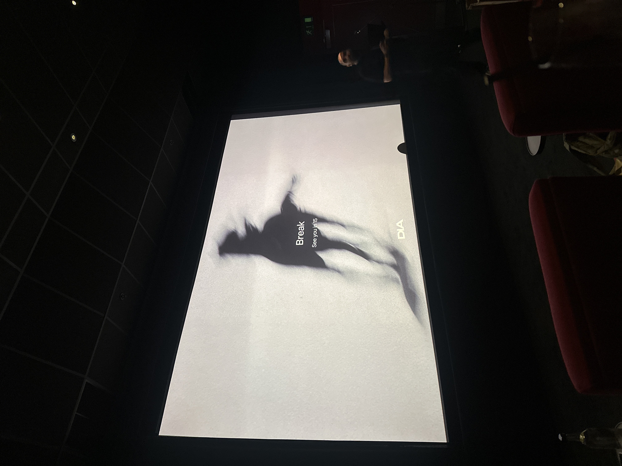 The image shows the interior of a cinema, possibly located in London, United Kingdom. A large screen displays a black and white advertisement or artwork featuring a human silhouette, and the words "Break" and "See you in 15." In the background, a person is standing taking a photo of the screen. Some dark red cinema seats are visible to the right. The walls are black with a gridded pattern.