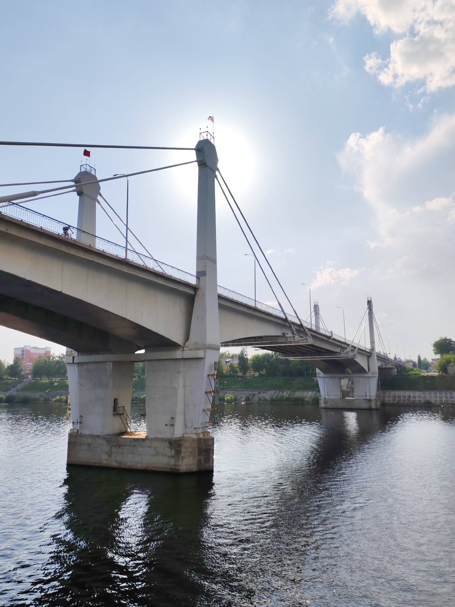 The image depicts a concrete bridge spanning a body of water, likely a river. The bridge has tall, prominent support structures that are situated in the water. The sun is positioned behind one of the bridge's support pillars, creating a bright glare. A red flag is visible on one of the pillars. The bridge is designed with a railing along its side, and a person is visible walking on the bridge. The water is calm with visible reflections of the bridge and sky. The background shows a cityscape with buildings and greenery along the riverbank. The location is identifiable as Kimry, Russia, known for its prominent bridge structures.