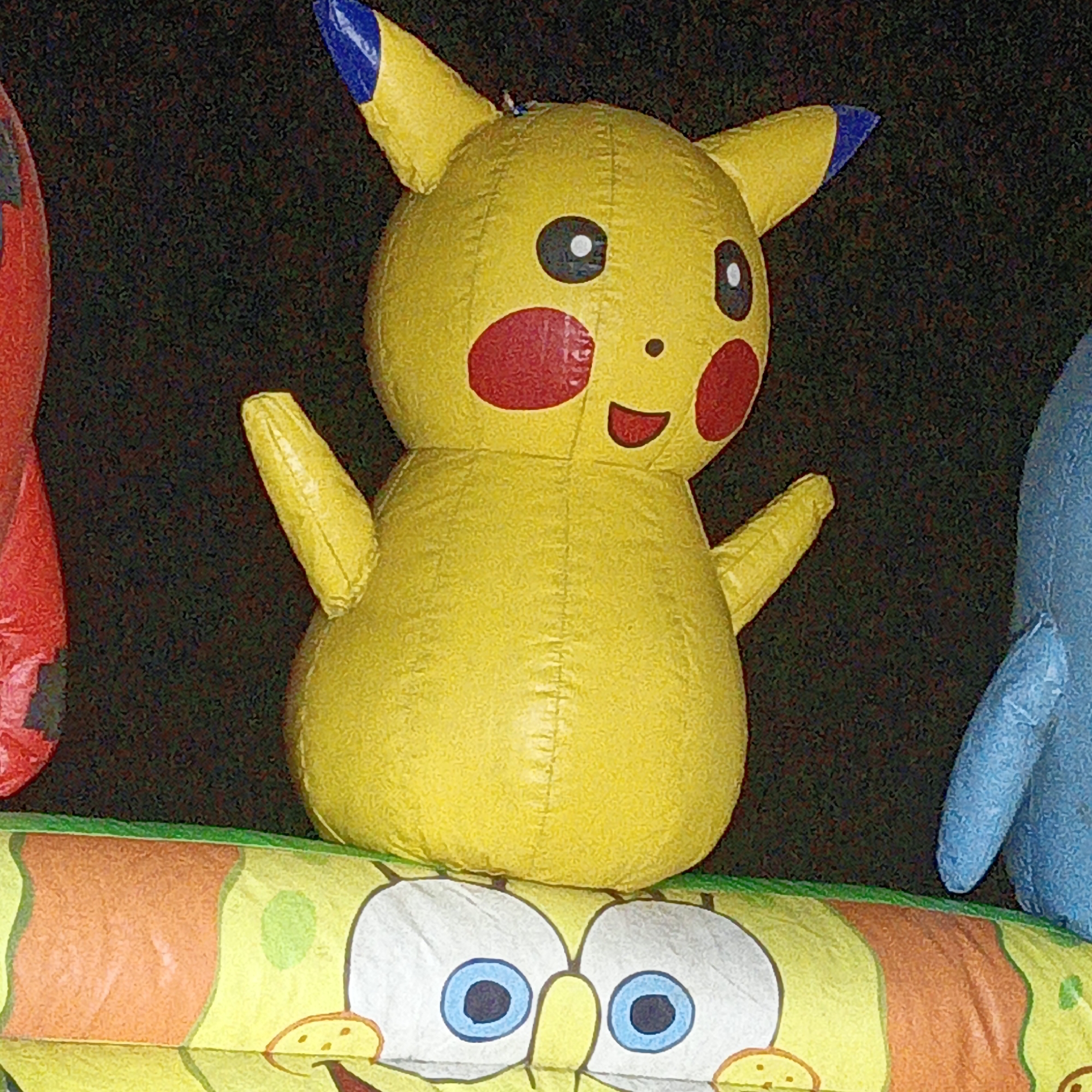 An inflatable Pikachu character, a popular Japanese anime character, is displayed outdoors. The Pikachu is yellow with red circular cheeks and blue-tipped ears. It is positioned in front of a dark, possibly night-time, background. Below the Pikachu, a portion of another inflatable object is visible, featuring a cartoonish face with large blue eyes and a yellow nose, resembling SpongeBob SquarePants. The image appears to be taken during a local event or celebration in Mangli, Indonesia, given the cultural context of such inflatable displays often seen during festivals or public gatherings. The overall scene suggests a festive or entertainment-oriented setting.