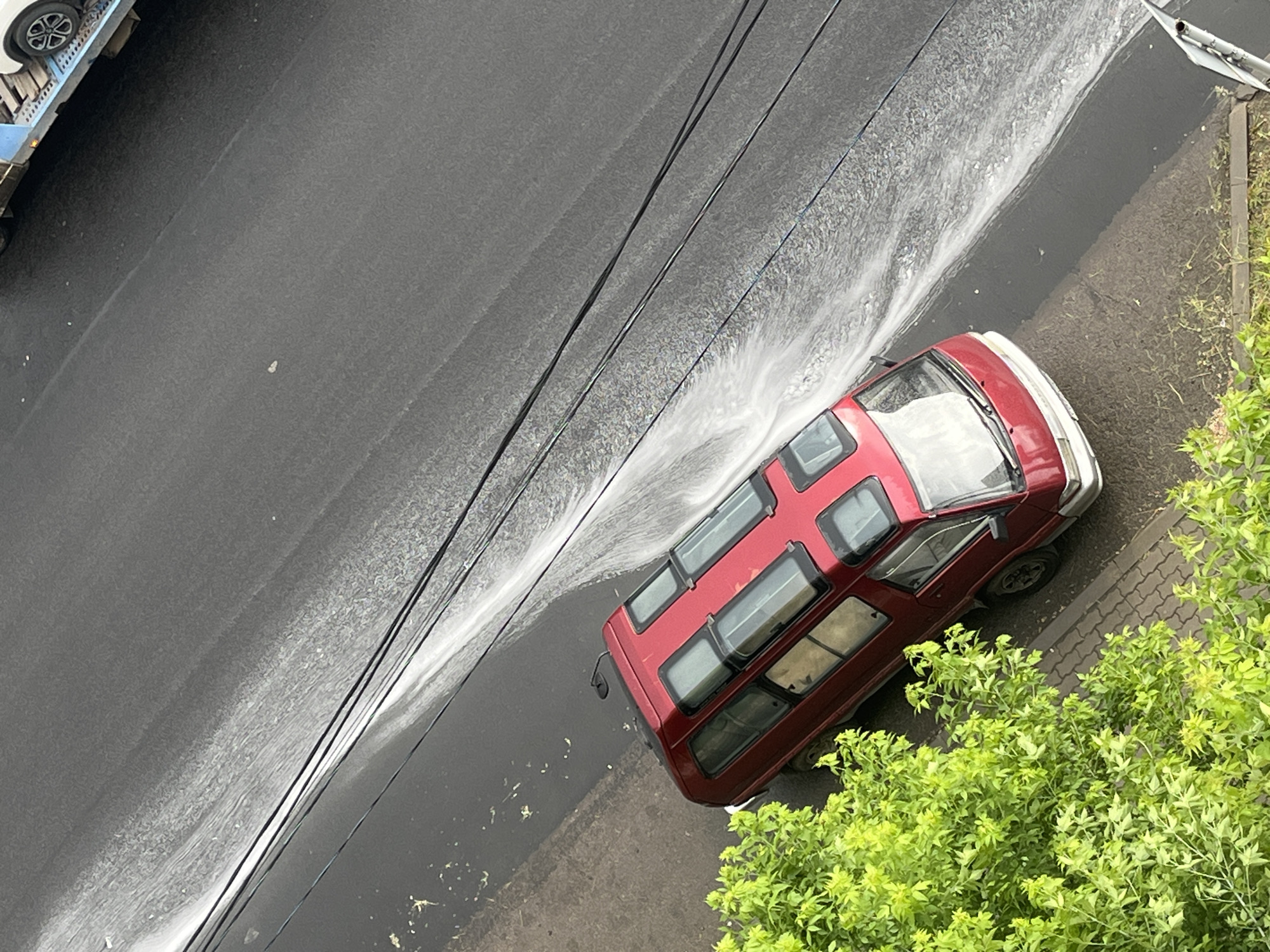 A red van is parked on an asphalt road next to a sidewalk lined with a green leafy tree. Water is spraying across the road from the right side of the image, creating a white foamy pattern on the dark asphalt. Three black electrical wires are stretched horizontally across the scene, above the van and the water spray. In the upper right corner, the rear of a white car on a blue trailer is visible, suggesting a street or parking area. The scene appears to be an outdoor environment, possibly a street in the city of Krasnoyarsk, Russia, given the context.