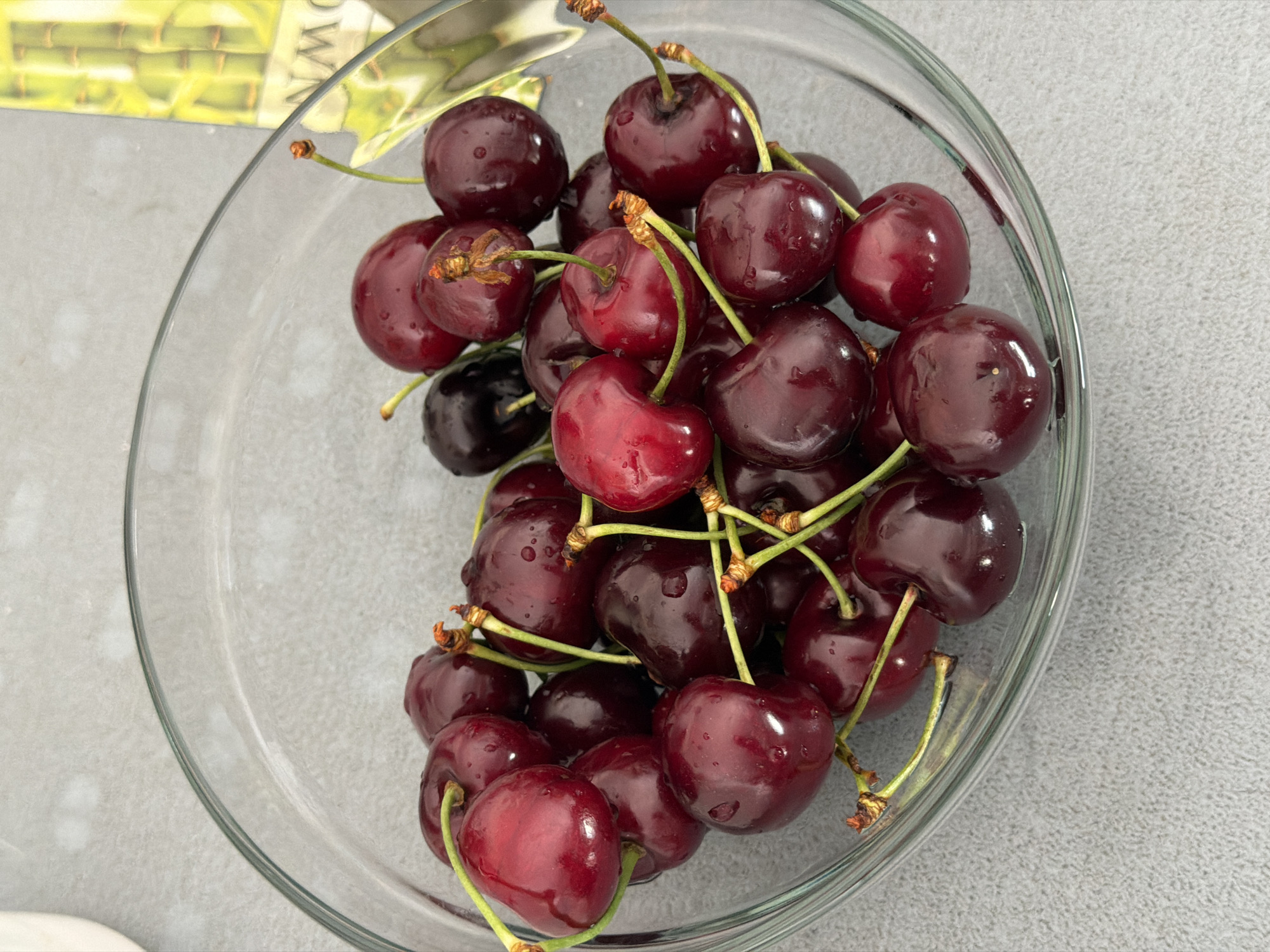 A clear glass bowl filled with dark red cherries sits on a light gray textured surface. The cherries are glossy and appear to be wet, with visible water droplets on their surfaces. Some of the cherries have long, green stems attached. In the background, out of focus, a portion of a decorative bamboo-themed item is visible on the right side. The image does not provide any information or visual cues to determine its location in the city of Burgas, Bulgaria.