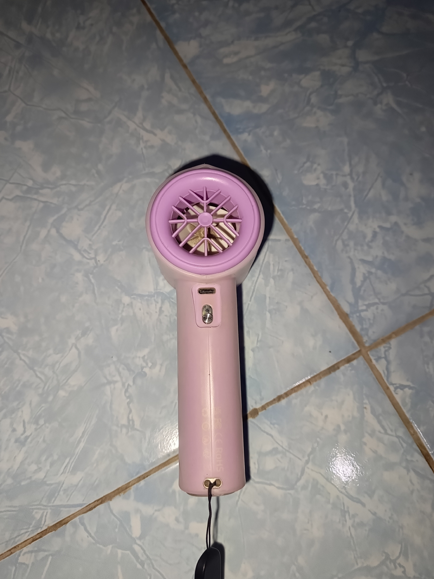 A pink handheld fan with a purple fan guard sits on a light blue tiled floor. The fan has a USB-C port and a power button located on the handle. A black lanyard is attached to the bottom of the fan. The floor is tiled with visible grout lines. The image appears to be taken indoors. There is no indication of the location being in Terbanggi Marga, Indonesia.
