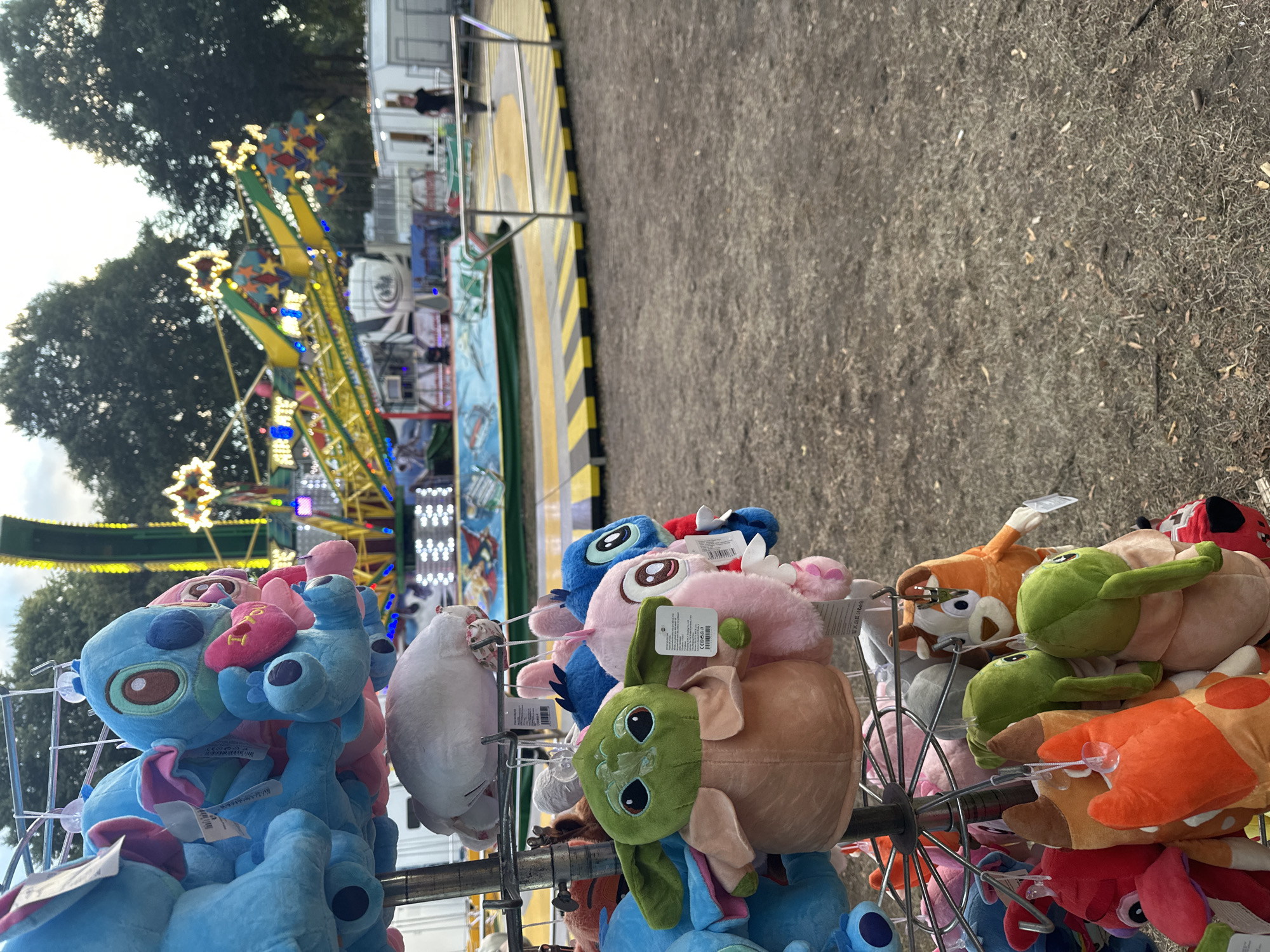 In Bruchhausen-Vilsen, Germany, a close-up shot captures a rack of assorted plush toys displayed outdoors, likely at a fairground or amusement park. The toys are varied, featuring popular characters like Stitch from Disney's Lilo & Stitch and Grogu (Baby Yoda) from The Mandalorian, along with other animal-themed plushies. The foreground is dominated by these colorful toys, some still bearing their tags. Behind them, a blurred background reveals a vibrant amusement park ride in operation, characterized by its bright lights and yellow and green structure. The ground appears to be a mix of dirt and dry grass. In the distance, a person is visible walking near a portable structure. The lighting suggests it is either late afternoon or early evening, with a soft, diffused light illuminating the scene.