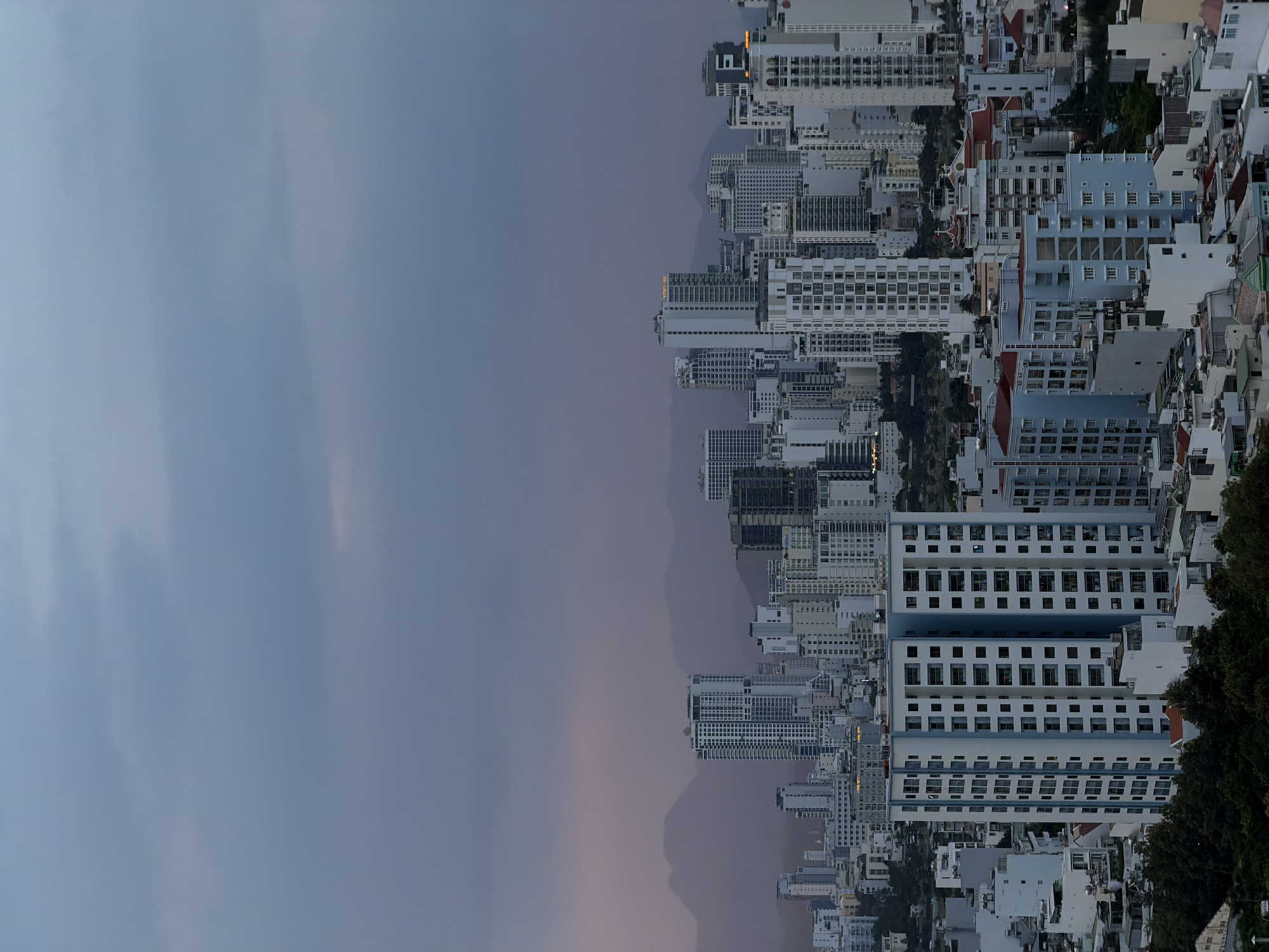 The image shows a cityscape of Nha Trang Ward, Vietnam, viewed from an elevated perspective at dusk. The scene is dominated by a dense collection of multi-story buildings, including many modern apartment complexes and hotels. The architecture is varied, with some buildings featuring a stark white facade and repetitive window patterns, while others have lighter blue accents or terracotta-tiled roofs.
The sky is overcast and a muted shade of blue and gray, suggesting either the approach of night or a cloudy weather condition. Faint hints of sunset colors, like a soft pink, are visible on the horizon, peeking through the atmospheric haze. In the background, the silhouettes of distant mountains can be seen, adding depth to the urban sprawl. On the right side of the image, a dense cluster of dark green trees marks the edge of the city or a natural landscape feature.
The overall mood is somewhat subdued, reflecting the dim light of dusk. There are no visible people in the frame, and the focus is entirely on the built environment and the natural elements that frame it. The image is oriented vertically, with the buildings appearing to extend downwards from the top of the frame. No discernible text is visible on any of the buildings or in the scene.