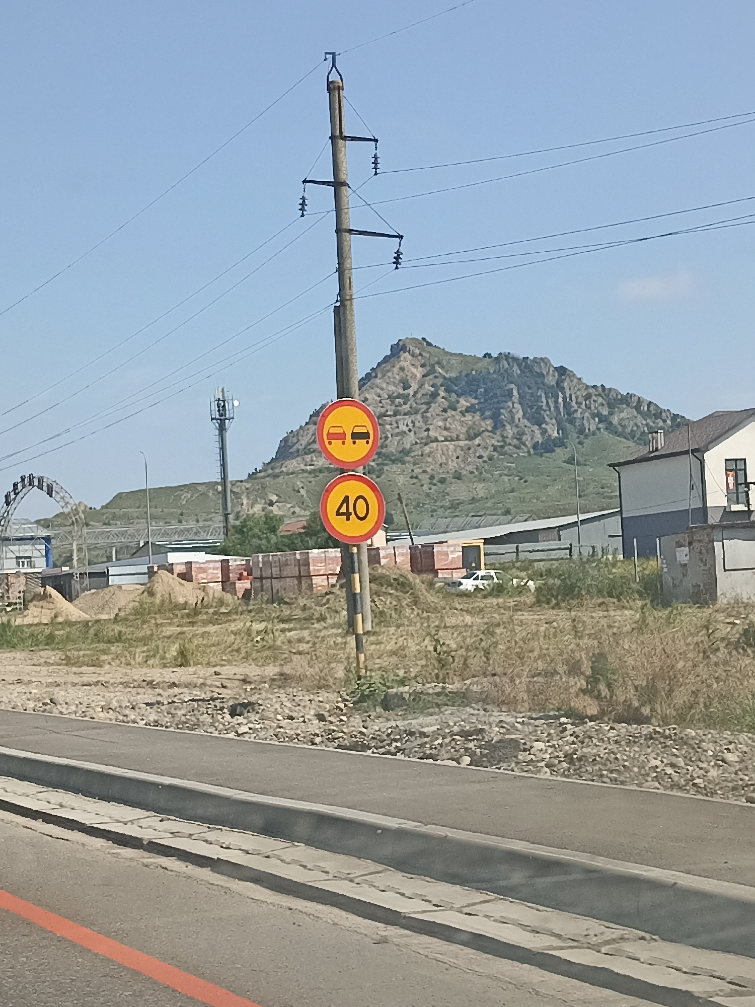 The image depicts a street scene in Vinsady, Russia, under a clear blue sky. A concrete utility pole stands prominently in the foreground, adorned with two circular traffic signs. The top sign features two red cars within a yellow circle, indicating a restriction on overtaking. Below it, another yellow circle contains the number "40," signifying a speed limit of 40 kilometers per hour.
The background reveals a rugged, rocky mountain range, partially covered with green vegetation. A modern building with a dark blue and white facade is visible to the right of the mountain. In the middle ground, construction materials, including piles of sand and stacks of bricks, are scattered across a dry, grassy area. A white vehicle is parked near a low-rise industrial-style building. A cellular tower with a ladder extends upwards in the distance, and a large arched structure, possibly part of a construction or entertainment facility, is situated to the left. The foreground shows a paved road with a gray curb and an orange line marking. The overall scene suggests a semi-urban or semi-rural environment undergoing development. There are no people visible in the image.
