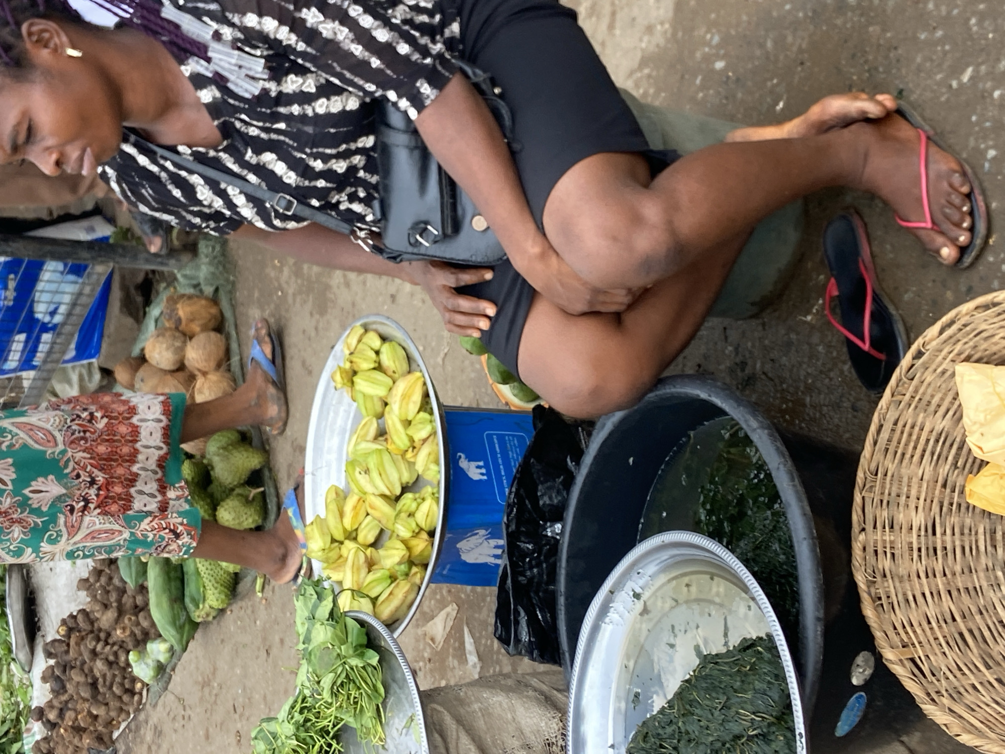 This is a medium shot, full-body photograph of a woman seated at a market stall in Nkpor, Nigeria. The woman is wearing a black and white striped short-sleeved blouse, a black skirt, and pink flip-flops. She has dark skin and dark braided hair with purple highlights. A black purse is slung over her shoulder. In front of her, on a blue scale, is a tray of star fruit. To her left, a large black basin filled with water and leafy green vegetables is visible, and to her right, a woven basket is partially visible. The ground appears to be wet and muddy. In the background, other market goods and people are present, suggesting a busy marketplace environment. The overall lighting indicates it is daytime, likely under overcast skies or in a shaded area.