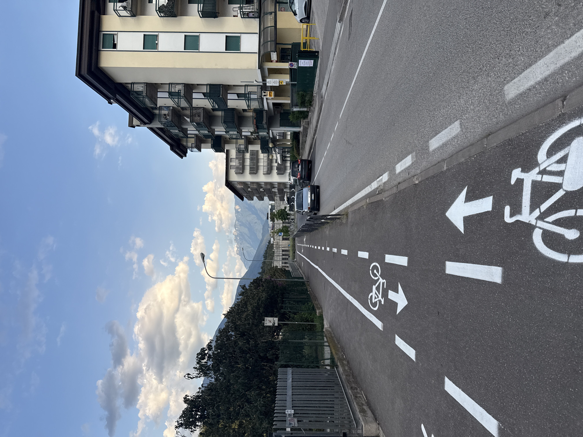 A view of a street in Trento, Italy, with a dedicated bicycle lane marked by white lines and bicycle symbols. The lane runs along the left side of the road, separated from the car traffic by a dashed white line. Several cars are driving on the road. On the right side of the road, there is a multi-story apartment building with balconies. Trees and mountains are visible in the background under a partly cloudy blue sky. The image appears to have been taken during the daytime.