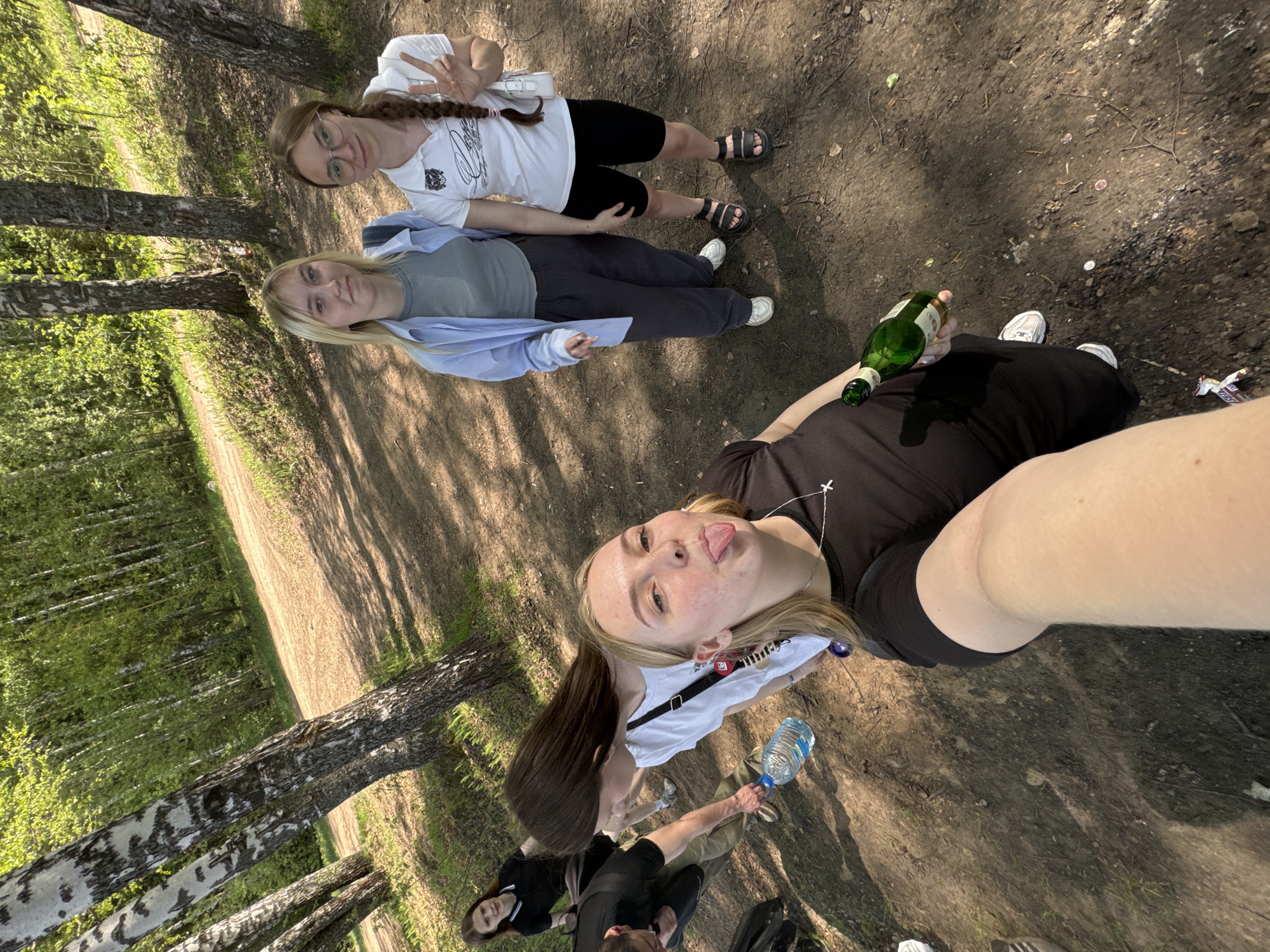 The image shows a group of young adults taking a selfie outdoors. The location appears to be a park or wooded area, judging by the trees and dirt ground. Three individuals are fully visible in the foreground, one holding a bottle and sticking their tongue out, likely taking the selfie. Another woman is lying down beside the selfie taker. Above them, a third woman is standing and making a peace sign. In the background, more individuals are visible, but their faces are less clear. One person is holding a water bottle. There is a birch forest in the background, which is a common landscape feature in Russia. While it's challenging to pinpoint the exact location without more context, considering that it’s taken in Perm, Russia, this could potentially be one of the city's parks or wooded areas along the Kama River. The ground has litter.