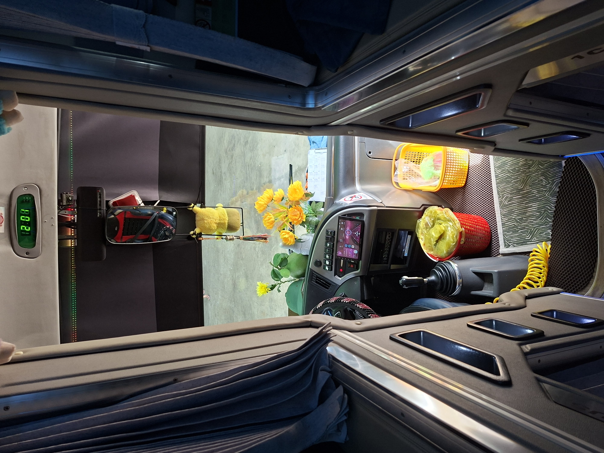 This is an interior view of a bus or a similar vehicle, likely taken in Tân Phú 2, Vietnam, looking towards the front cab. The viewpoint is from the aisle, with dark grey or black molded paneling on either side. On the left, a pleated blue-grey curtain is drawn. The front of the bus features a dashboard with a digital clock displaying "2:01", various buttons and switches, a touchscreen display showing a map, and a steering wheel adorned with red and black beads.
Objects on the dashboard include a green hat, a green ornamental gourd, and a vase with artificial yellow flowers. To the right of the steering wheel, there's a red basket filled with yellow plastic bags, and a smaller yellow plastic basket containing some items. A coiled yellow cord is visible near the gear shift. The floor is covered with textured mats. The overall impression is of a well-maintained and somewhat personalized interior, with a mix of functional elements and decorative items. The background appears to be a concrete floor, possibly a garage or parking area.