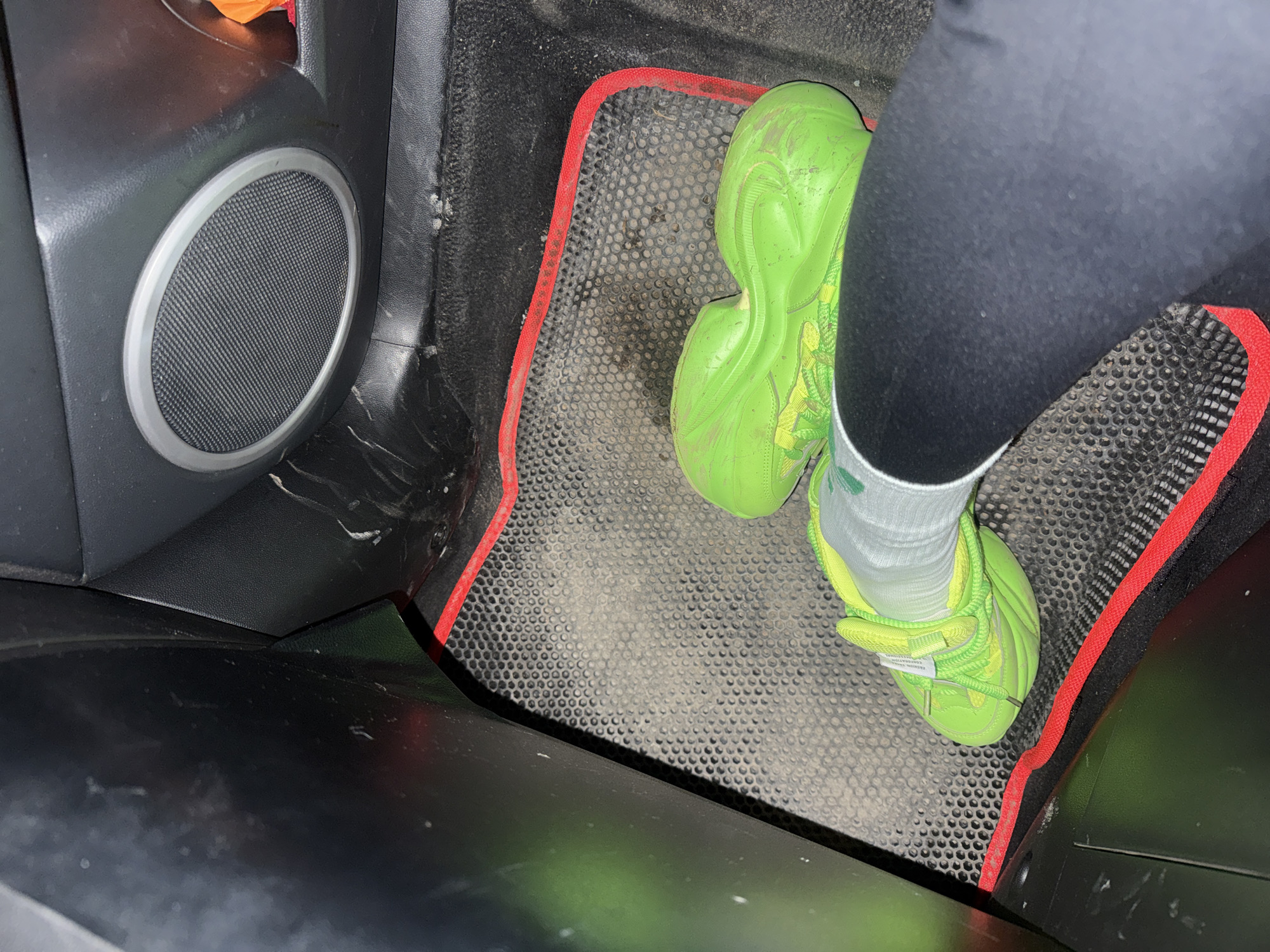 The image shows the interior of a vehicle, likely in the driver's side footwell. A person wearing black leggings and white socks is present, with their foot resting on a black rubber floor mat with a red border. The person is wearing bright green sneakers. To the left, a portion of the car's dashboard is visible, featuring a circular speaker. The environment shows a dirty and well-used interior. There is no information to suggest the picture was taken in the city of Kauchuk Stantsiya, Russia.