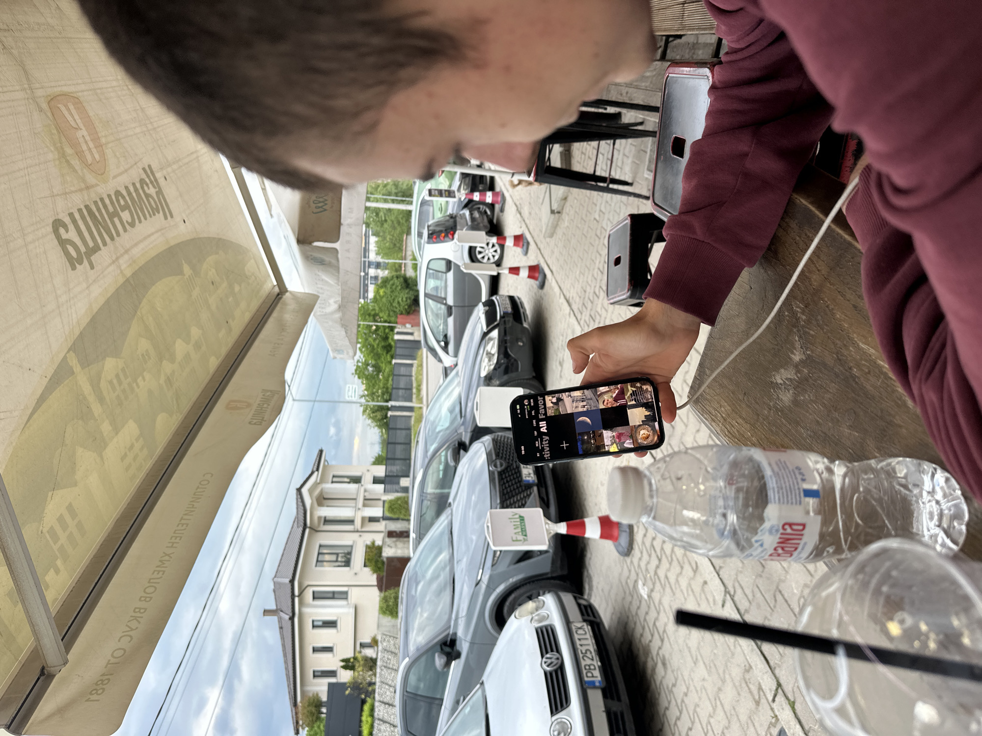 A close-up shot presents a person holding a smartphone displaying what appears to be a social media interface. Adjacent to the phone, a water bottle with the brand "Bania" is visible on a wooden table. The environment is an outdoor cafe, characterized by umbrellas and chairs. In the background, a parking area filled with various cars is noticeable, bordered by buildings. The license plate number of the nearest vehicle is "PB 2511 CK". A sign with the word "FAMILY" is attached to a safety cone. The image composition suggests the location is in a city, likely Plovdiv, Bulgaria, based on the license plate and the local brand of bottled water.