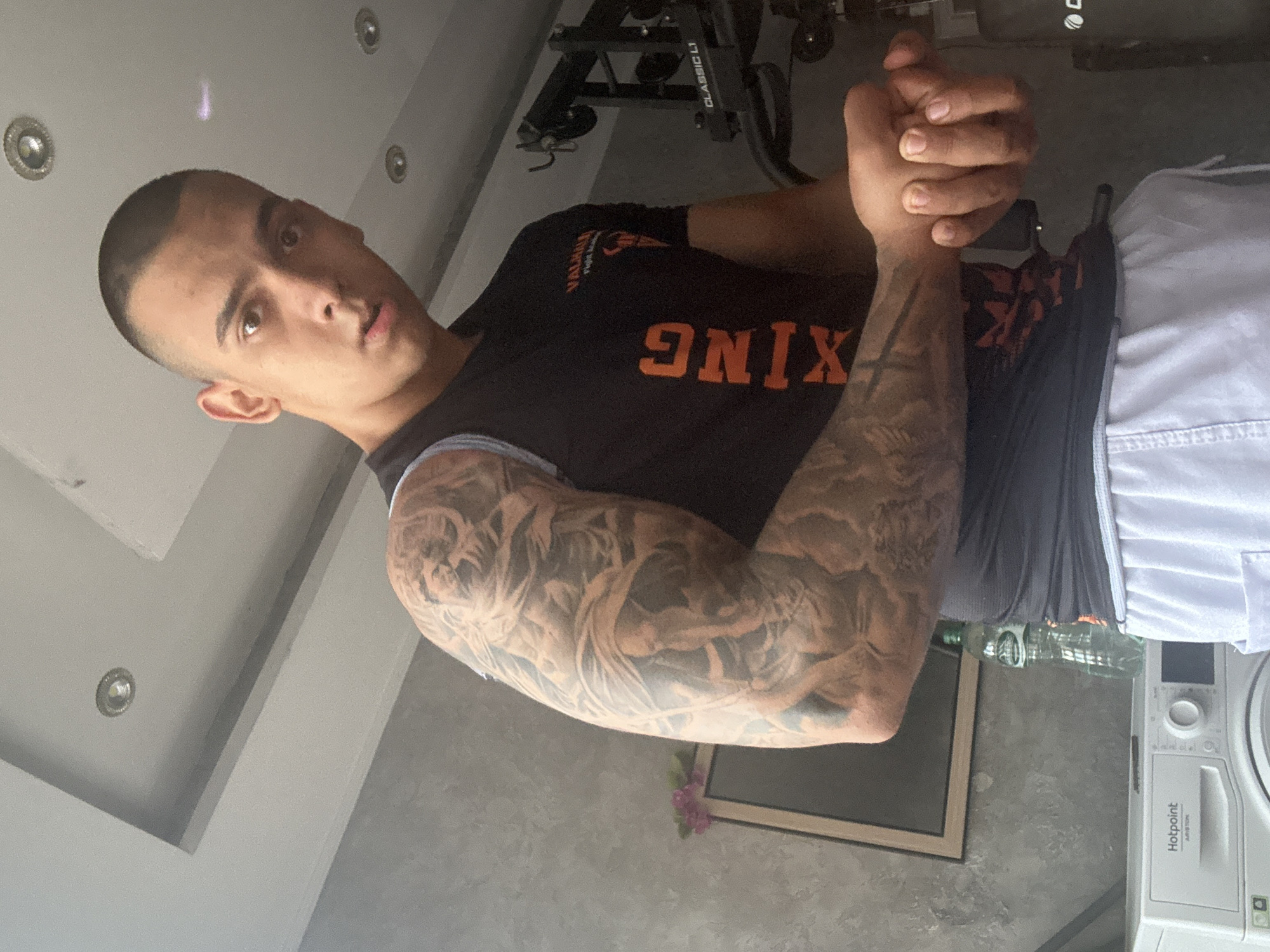 A man with a closely shaved haircut is standing in an indoor setting. He has extensive tattoos covering his left arm. He is wearing a black t-shirt with orange lettering that reads "ONIX," partially obscured, and the word "VALHALLA" is visible above it. He also appears to be wearing shorts with a dark waistband, overlaid by white fabric. His hands are clasped together in front of him.
In the background, a ceiling with recessed lighting can be seen, along with a piece of gym equipment, possibly a treadmill or exercise machine. A picture frame is on the wall in the middleground and a white appliance, possibly a washing machine with the brand name "Hotpoint," is also visible. The location is Unknown, Unknown.