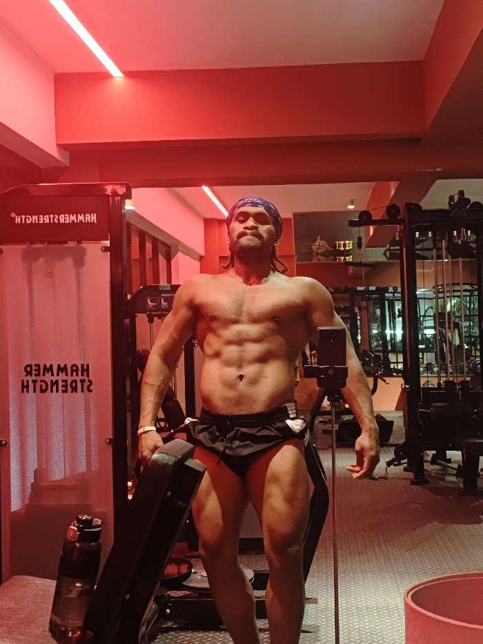 A male individual with a muscular physique is depicted in a full-length, front-facing photograph within a gymnasium in the Chongqing High-tech Zone, China. The subject, shirtless and wearing black athletic shorts with a blue patterned bandana, stands with his head slightly elevated. His left hand grips a black padded component, consistent with a segment of a weight bench, which rests against his upper left thigh. A smartphone, secured on a black tripod, is positioned to the subject's immediate right (viewer's left) for image capture. The gym interior features red ambient lighting emanating from ceiling-mounted linear fixtures, illuminating textured floor tiling. Reflected in a large mirror on the left side of the frame, the inverted text "HAMMER STRENGTH" is visible on a piece of equipment. Additional exercise machinery and a dark water bottle are present in the background.