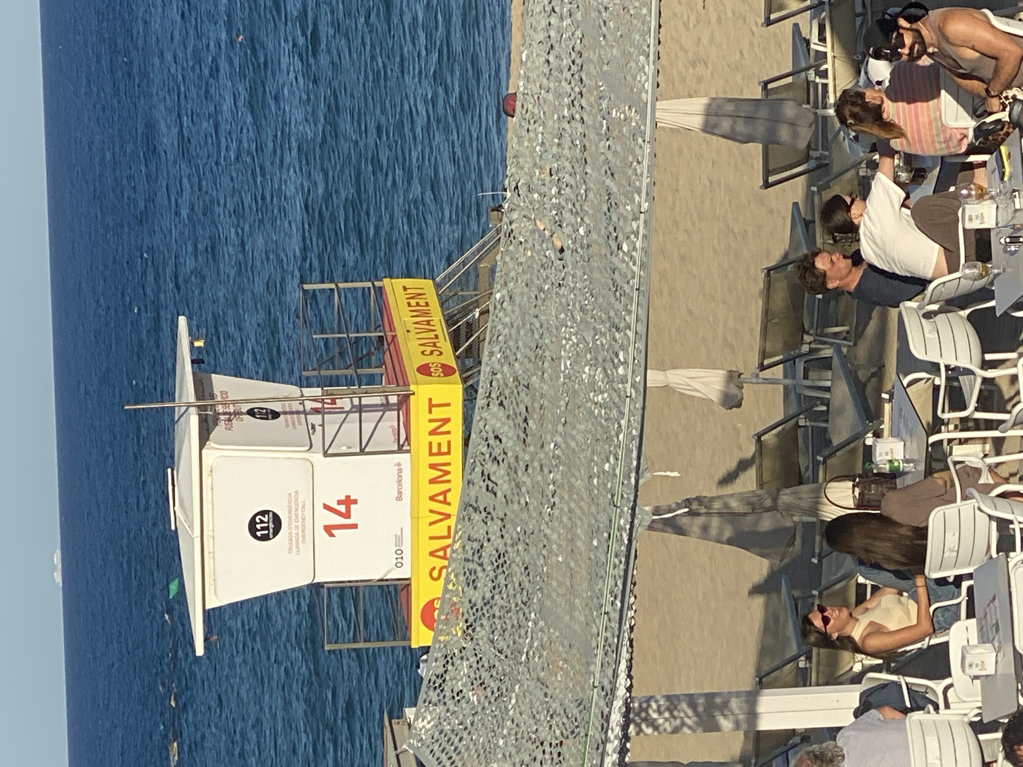 This image captures a vibrant, sunny beach scene in Barcelona, Spain, featuring a lifeguard tower and an adjacent beachfront restaurant. On the left, the calm, deep blue Mediterranean Sea extends to the horizon under a clear sky. A prominent white lifeguard tower, marked with a large red "14," stands near the water. It displays emergency contact information: "112 TRUCADA D'EMERGENCIA LLAMADA DE EMERGENCIA EMERGENCY CALL," and a yellow sign with "SOS SALVAMENT" and "010 Barcelona."
To the right, a sandy beach area gives way to an outdoor restaurant, where numerous individuals are seated at tables with white chairs under umbrellas. Patrons, including a bearded man in a tank top, a woman in a striped shirt, and another man in a white shirt with sunglasses, are seen relaxing and enjoying drinks, some looking towards the ocean. In the foreground, a woman in a light-colored top with sunglasses is smiling. The atmosphere is relaxed and social, typical of a daytime outing. Bright sunlight casts clear shadows, indicating a clear, sunny day. A mesh screen acts as a partition between the restaurant and the immediate beach area.