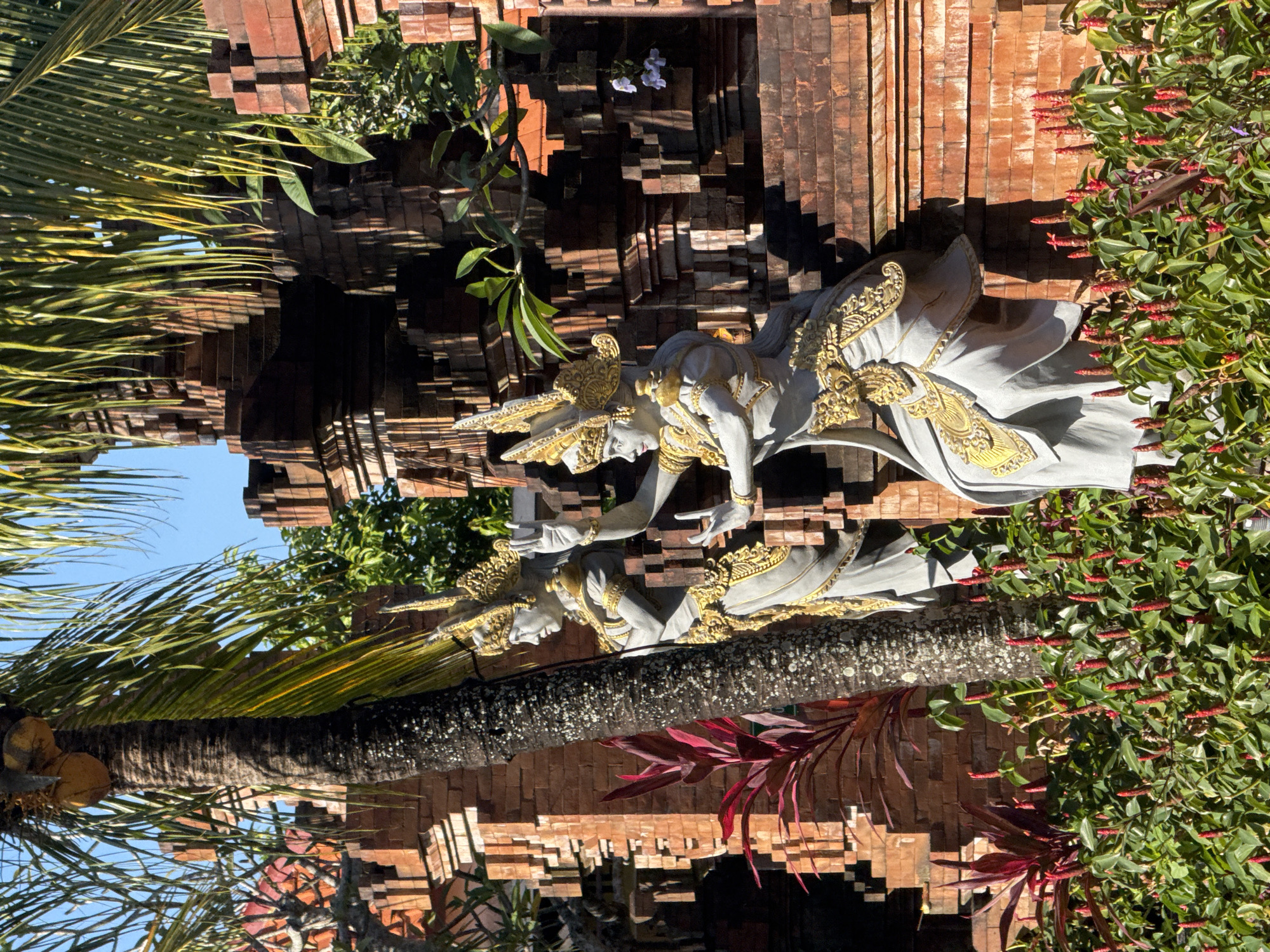 The image depicts a pair of statues in Kutuh, Indonesia, positioned against a tiered brick structure. The statues appear to be Balinese in style, featuring ornate gold embellishments. A palm tree is seen on the left, and flowering bushes with red blossoms are visible on the right. The background suggests an outdoor, potentially garden-like environment under a clear sky.