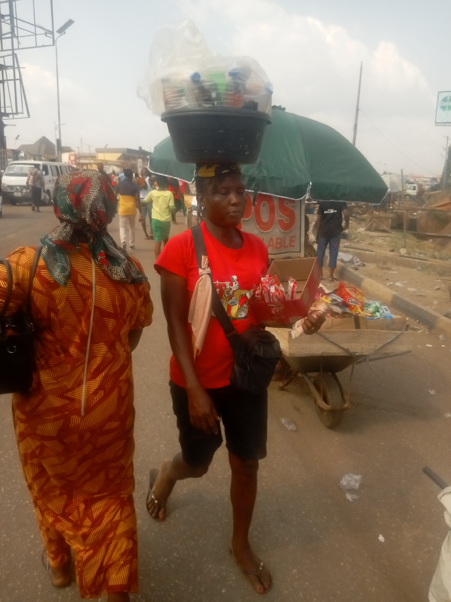 The image depicts a bustling street scene in Onitsha, Nigeria. In the foreground, a woman walks toward the left, wearing a red short-sleeved top, black shorts, and dark flip-flops. She balances a black basin filled with plastic-wrapped items on her head and carries a black crossbody bag. In her right hand, she holds a red cardboard display box containing individually wrapped snacks, and several Nigerian Naira banknotes are visible in her left hand. To her left and slightly behind her, another woman walks away from the camera, wearing a long, patterned orange and yellow dress and a colorful headscarf, with a black handbag on her left shoulder.
To the right of the central woman, a green market umbrella provides shade over a makeshift stall. A white sign with red lettering reading "POS ABLE" is partially visible beneath the umbrella. A wooden wheelbarrow with a single wheel serves as a display stand, laden with various packaged snacks, including "Gala" branded items. The ground beneath the wheelbarrow and extending into the background is an unpaved, dusty area with visible litter.
In the mid-ground and background, numerous people are walking. A white van or bus is visible on the left side of the street, with other vehicles further back. Street infrastructure includes a tall light pole and a metal frame structure on the left. The sky is bright and appears overcast or hazy. A distant billboard with partial "Dettol" branding is visible on the far right. The street itself is paved asphalt, showing signs of wear and scattered debris.
