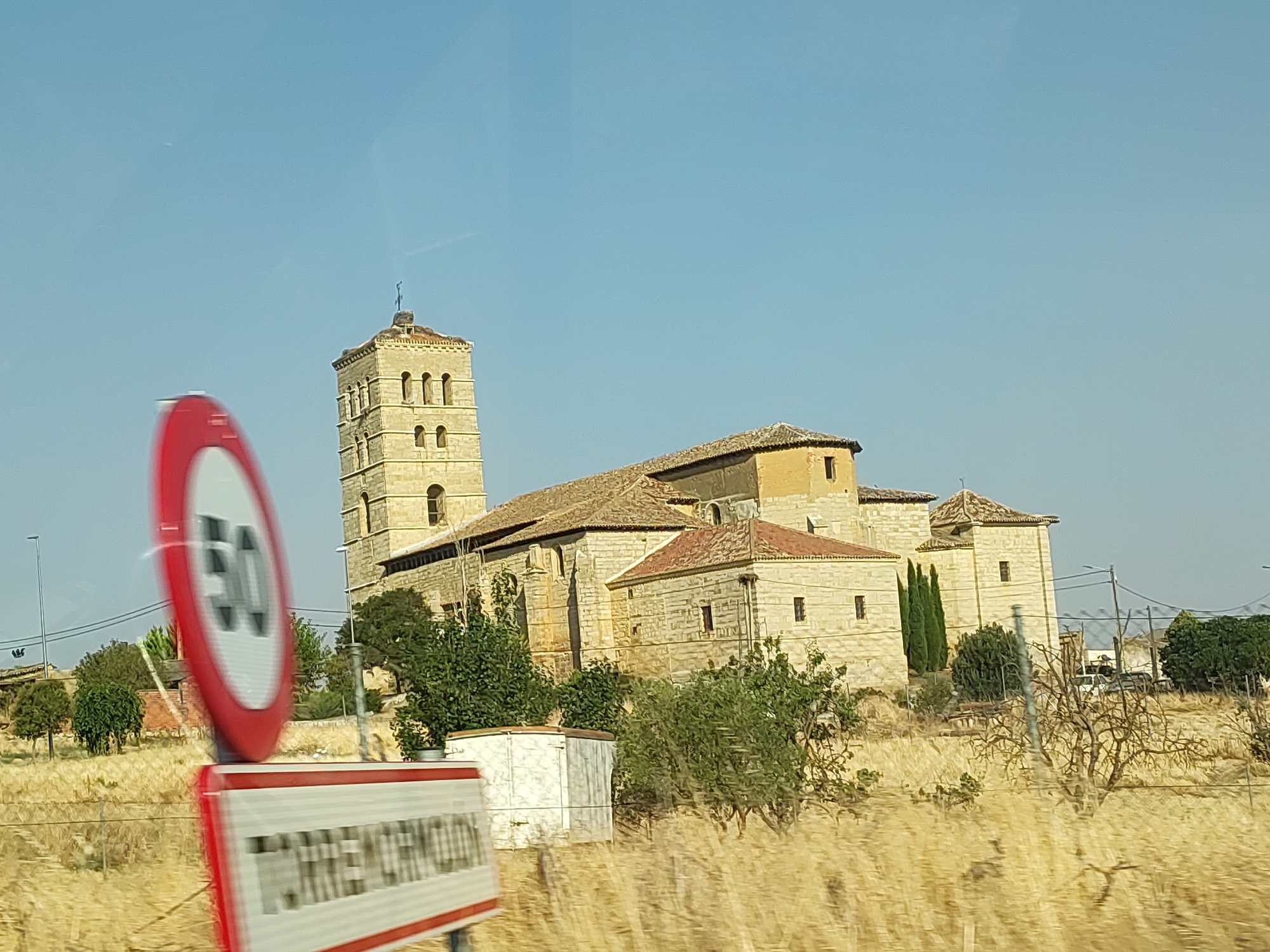 The image captures a sunny day scene in Torremormojón, Spain, featuring a stone church with a prominent bell tower. A red and white circular speed limit sign, indicating 50, is prominently displayed in the foreground, partially obscuring a rectangular sign with white text on a red and white background. The church, constructed of light-colored stone, has a tiled roof and multiple windows. The surrounding environment consists of dry, golden grass, scattered trees, and some small buildings. The sky is clear and blue. The image appears to have been taken from a moving vehicle due to the motion blur on the signs.