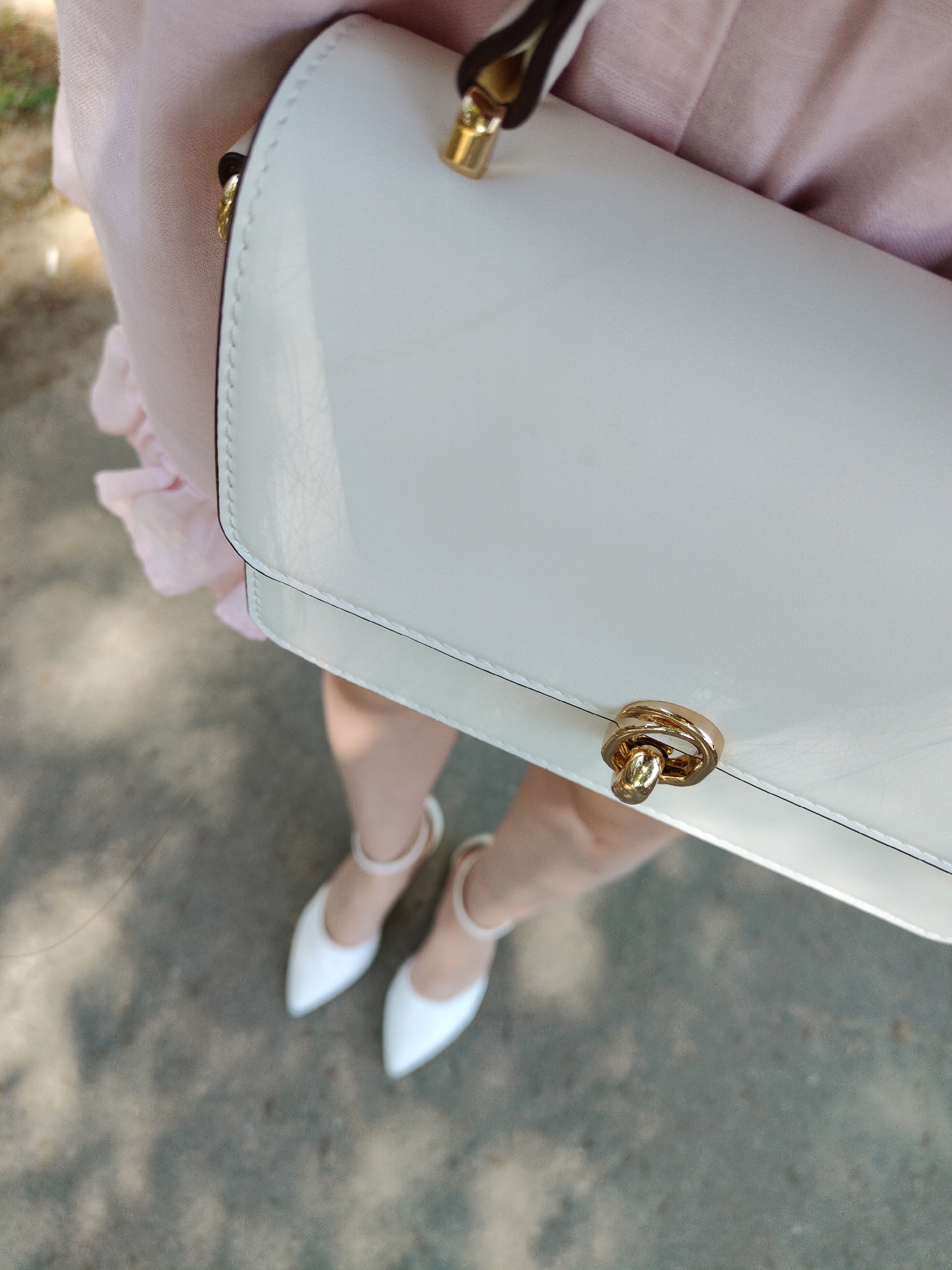 A top-down medium shot depicts a person holding a white handbag with gold hardware, wearing a light pink garment, and matching white shoes with ankle straps. They are standing on a concrete surface. The location is not specified in the image.