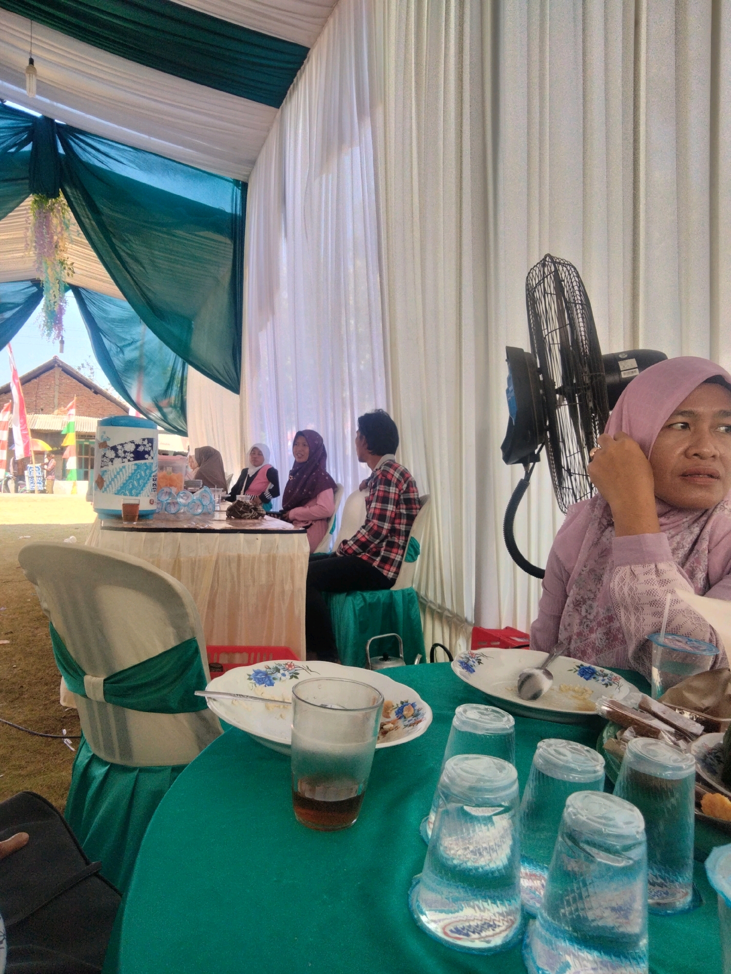 The image depicts an outdoor event under a tent, likely a reception or gathering, in Cilacap, Indonesia. In the foreground, a teal-colored tablecloth covers a round table laden with several plastic cups, some filled with a clear liquid and others empty. A glass of iced tea sits on the table, next to a plate with a floral pattern and a spoon. A woman in a lavender hijab and matching outfit is seated at the table, looking to her right, her hand near her face. Behind her, a large black fan is mounted on a stand. Further back, several other individuals are seated at tables, including two women wearing hijabs and a man in a red and white plaid shirt. The tent is decorated with draped green and white fabric. In the background, a building with red-tiled roofs and flags is visible, suggesting the event is taking place near a residential or community area. The ground appears to be covered in dry grass or dirt.