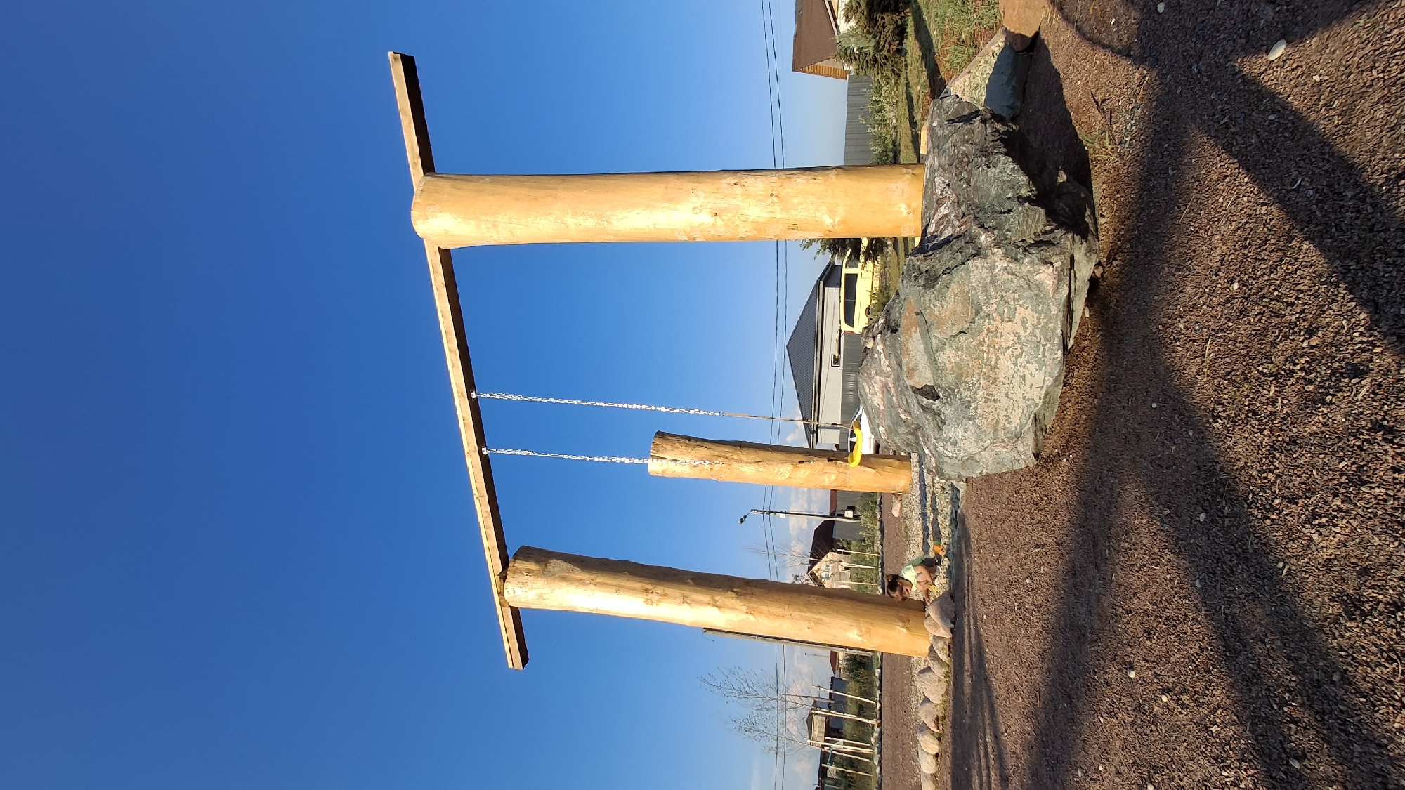 In Timmolovo, Russia, a unique swing set constructed from three large, rough-hewn wooden logs supports a wooden beam from which hang two metal chains. One chain has a yellow swing seat attached, while the other appears to be empty. In the foreground, a large, dark gray boulder with streaks of green and pink rests on reddish-brown gravel. A small child, wearing a light-colored shirt and orange shoes, is seated on the ground near the base of one of the wooden supports, playing with small stones. The area around the swing set is also covered in gravel. In the background, a grey metal fence encloses a grassy area, beyond which are several houses with dark roofs and a yellow minivan. The scene is illuminated by bright sunlight against a clear blue sky.