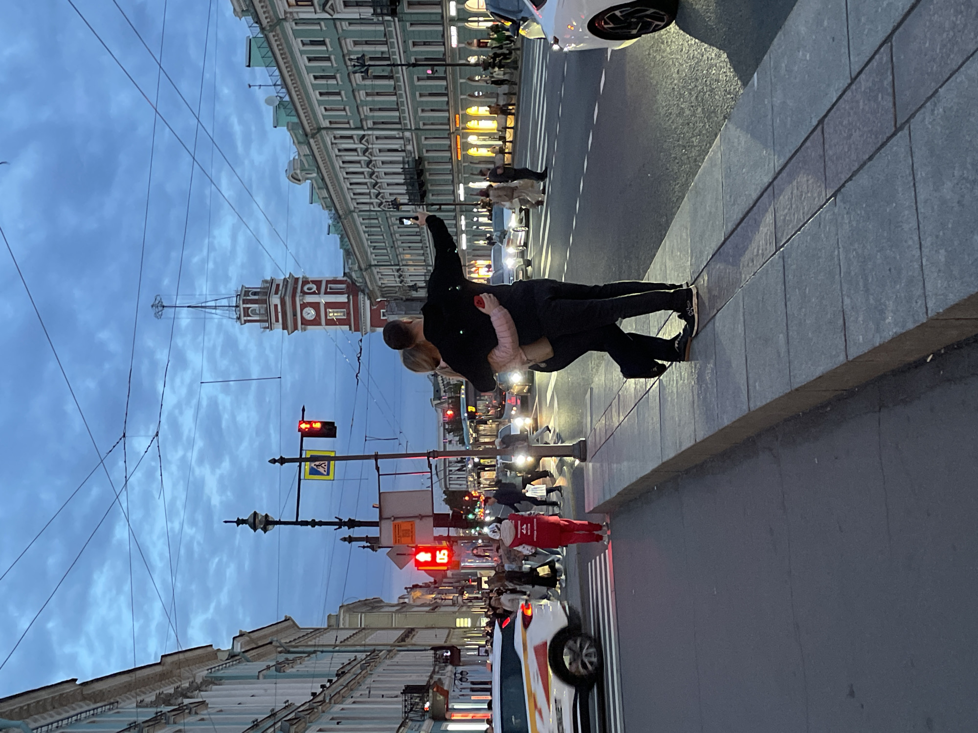 The image depicts a street scene in Saint Petersburg, Russia, at dusk. The sky is a deep blue with scattered clouds, and the streetlights and building lights are illuminated.
In the foreground, a couple is standing on a sidewalk, with the man taking a selfie with his phone. The woman is leaning into him. They are dressed in casual clothing.
The street is busy with traffic, including cars and a bus. Pedestrians are crossing the street at a marked crosswalk. A person in a red vest and pants is standing at the crosswalk, facing away from the camera.
The buildings lining the street are tall and appear to be in an architectural style common to Saint Petersburg, with ornate facades and many windows. A prominent clock tower with a red base and white top is visible in the background. The overall impression is of a lively urban environment at the end of the day.