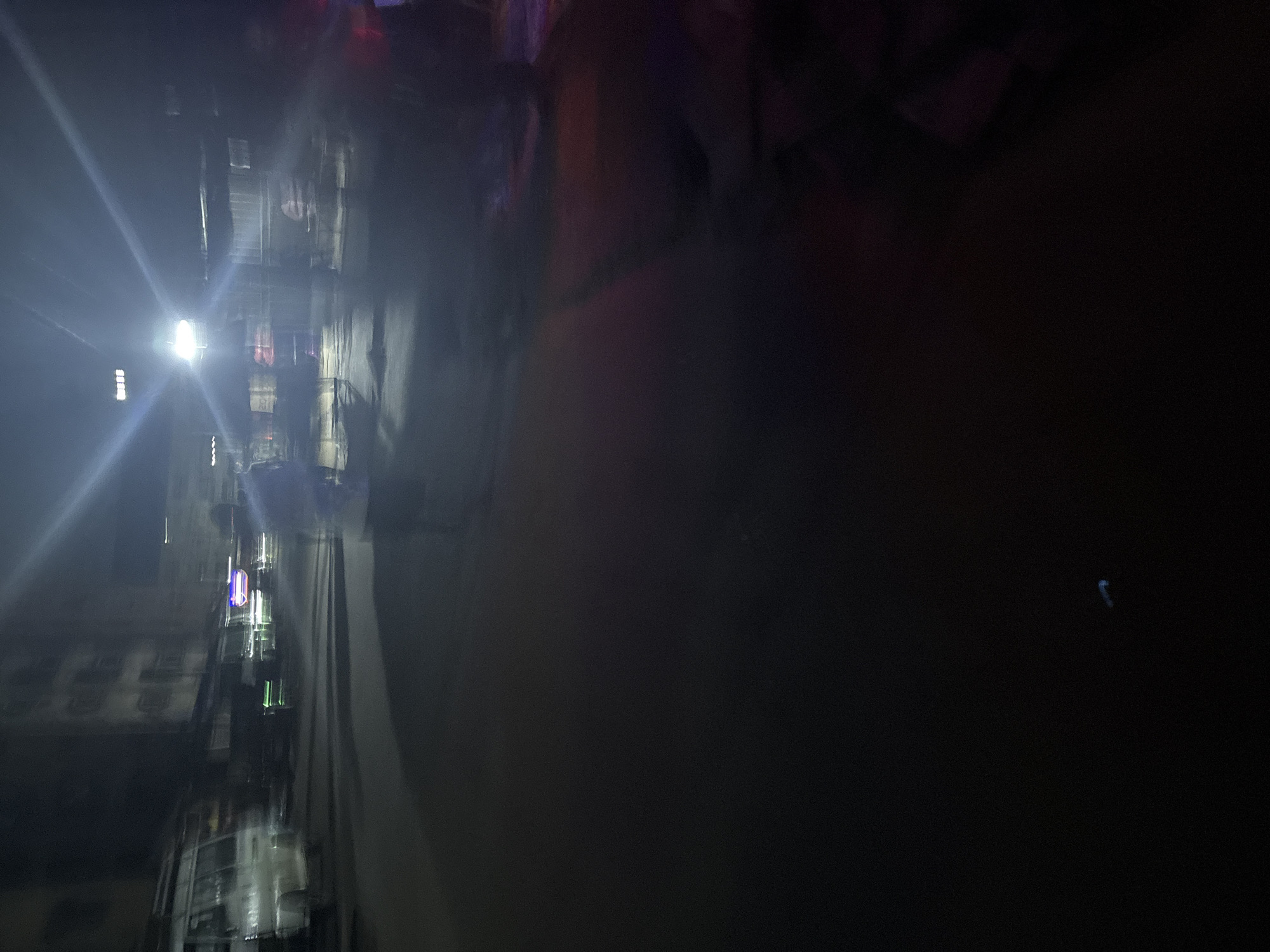 The image depicts a nighttime scene on a street in Onitsha, Nigeria. The street is paved and appears to be moderately lit, with a bright light source in the upper center casting lens flares. Several vehicles are visible, including a white van on the left side of the frame, which is partially obscured and appears to be moving. There are also blurred figures of people in the background, some of whom appear to be standing or walking. Structures, possibly shops or buildings, line the street. The overall impression is one of a busy urban environment at night, with a sense of motion and activity. The ground is dark and reflective in places, suggesting a wet surface or low light conditions. The image is characterized by motion blur, especially in the foreground and mid-ground, indicating that the photograph was likely taken from a moving vehicle or with a slow shutter speed.