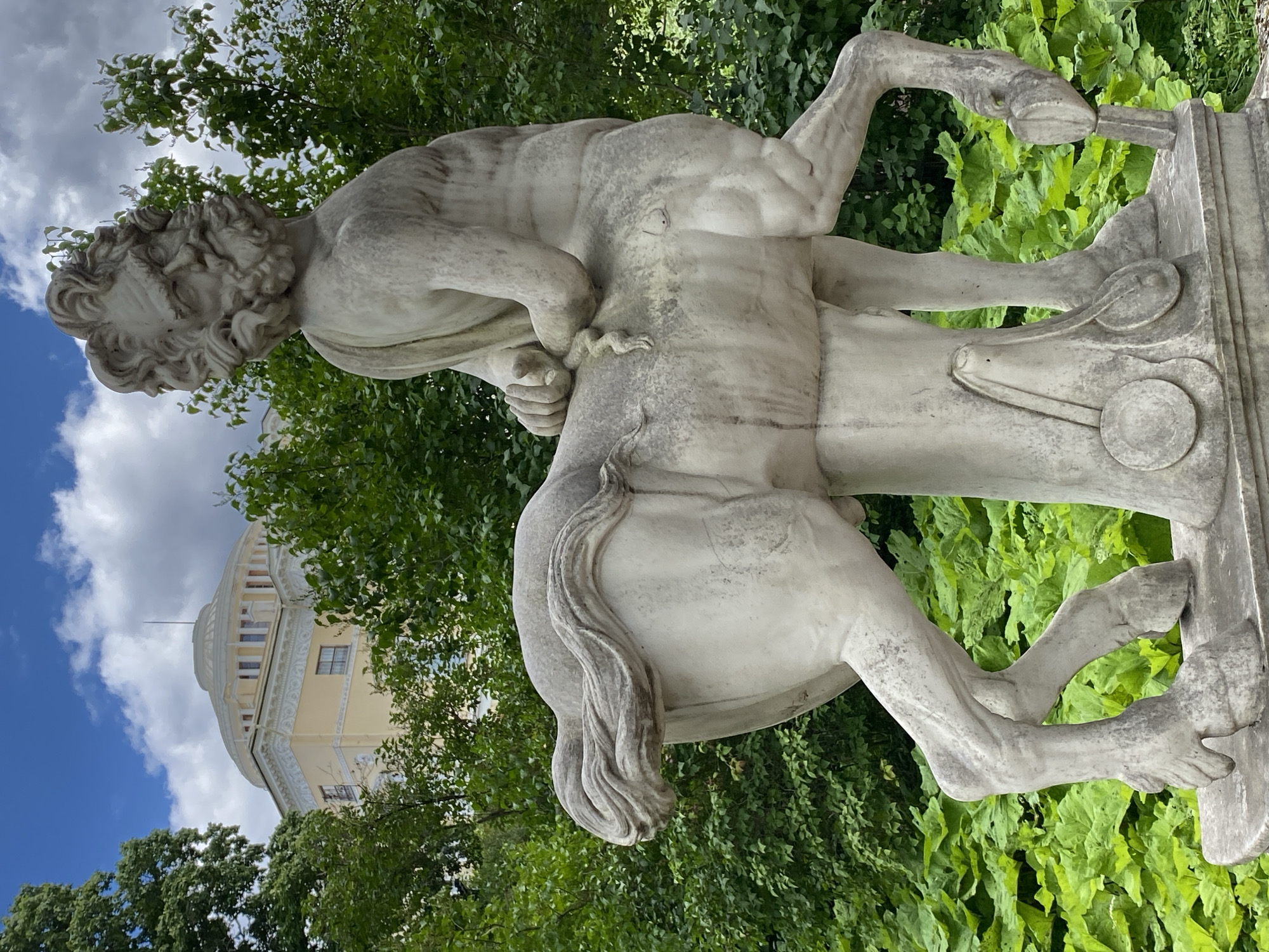 The image shows a stone sculpture of a centaur in the Pavlovsk Park, located in the city of Pavlovsk, Russia. The centaur sculpture is depicted in marble, with the upper body of a man and the lower body of a horse. It is positioned on a stone pedestal surrounded by lush green foliage. In the background, a neoclassical building with a domed roof is visible, partially obscured by trees. The sky is partly cloudy.