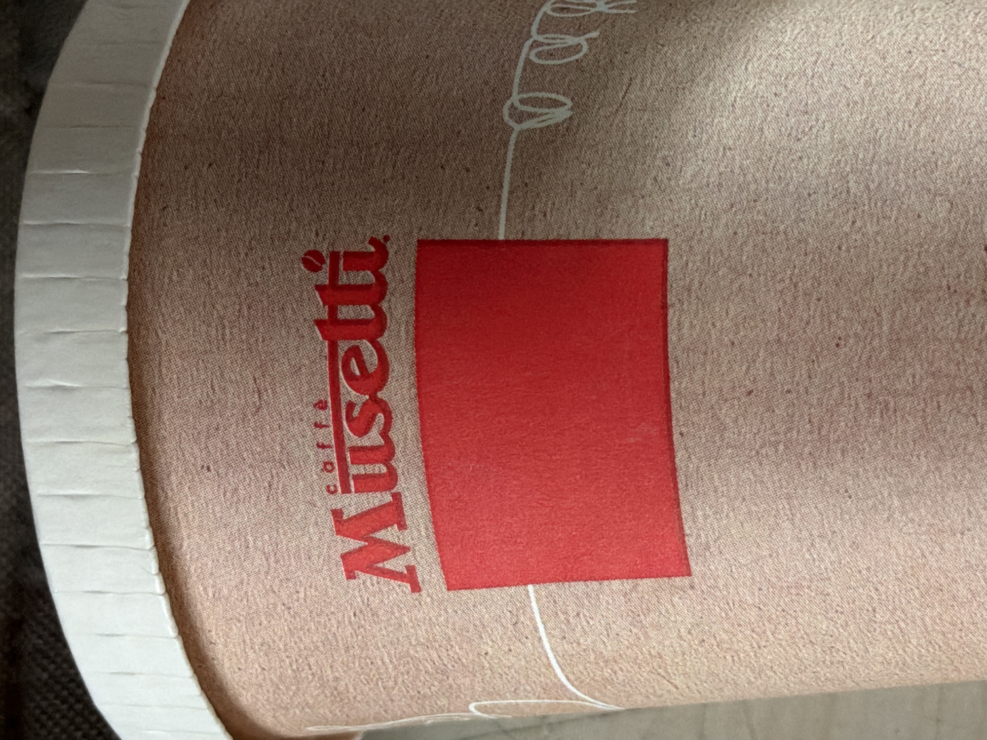 The image is a close-up of a paper cup with the "Musetti caffè" logo printed in red. A solid red rectangle is also printed on the cup. The cup has a white, ribbed rim. The background is dark and out of focus, suggesting it is likely a disposable coffee cup intended for on-the-go consumption, as might be found in various establishments in Piacenza, Italy.