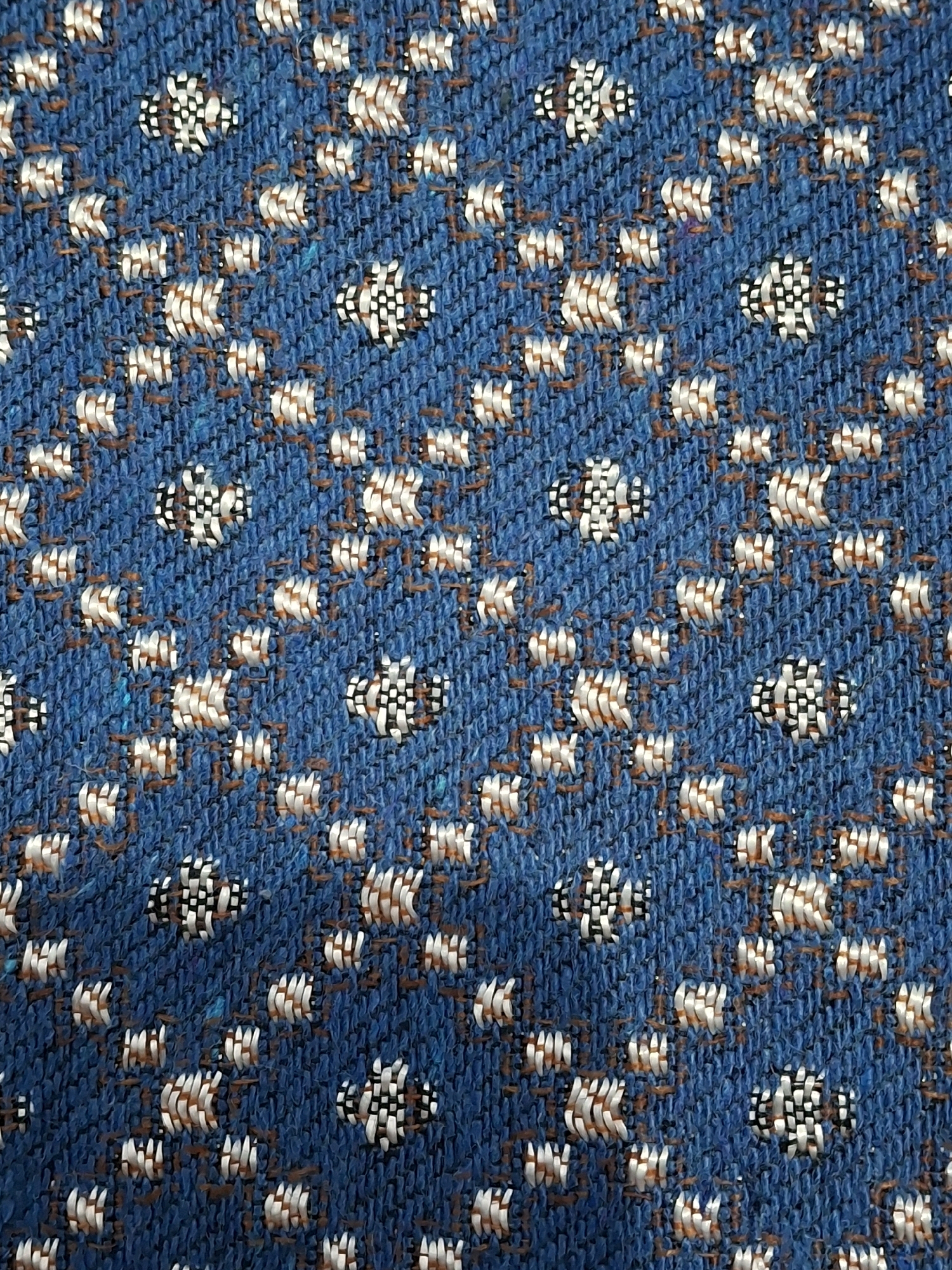 The image shows a close-up, top-down view of a blue textured fabric with a repeating pattern of small, abstract white and black designs. The fabric appears to be a woven material with a slightly raised texture. The pattern is somewhat scattered and irregular, with individual elements appearing in clusters of varying densities. The overall impression is of a decorative textile, possibly upholstery or a garment. There is no information within the image to suggest a location in Lumajang, Indonesia.
