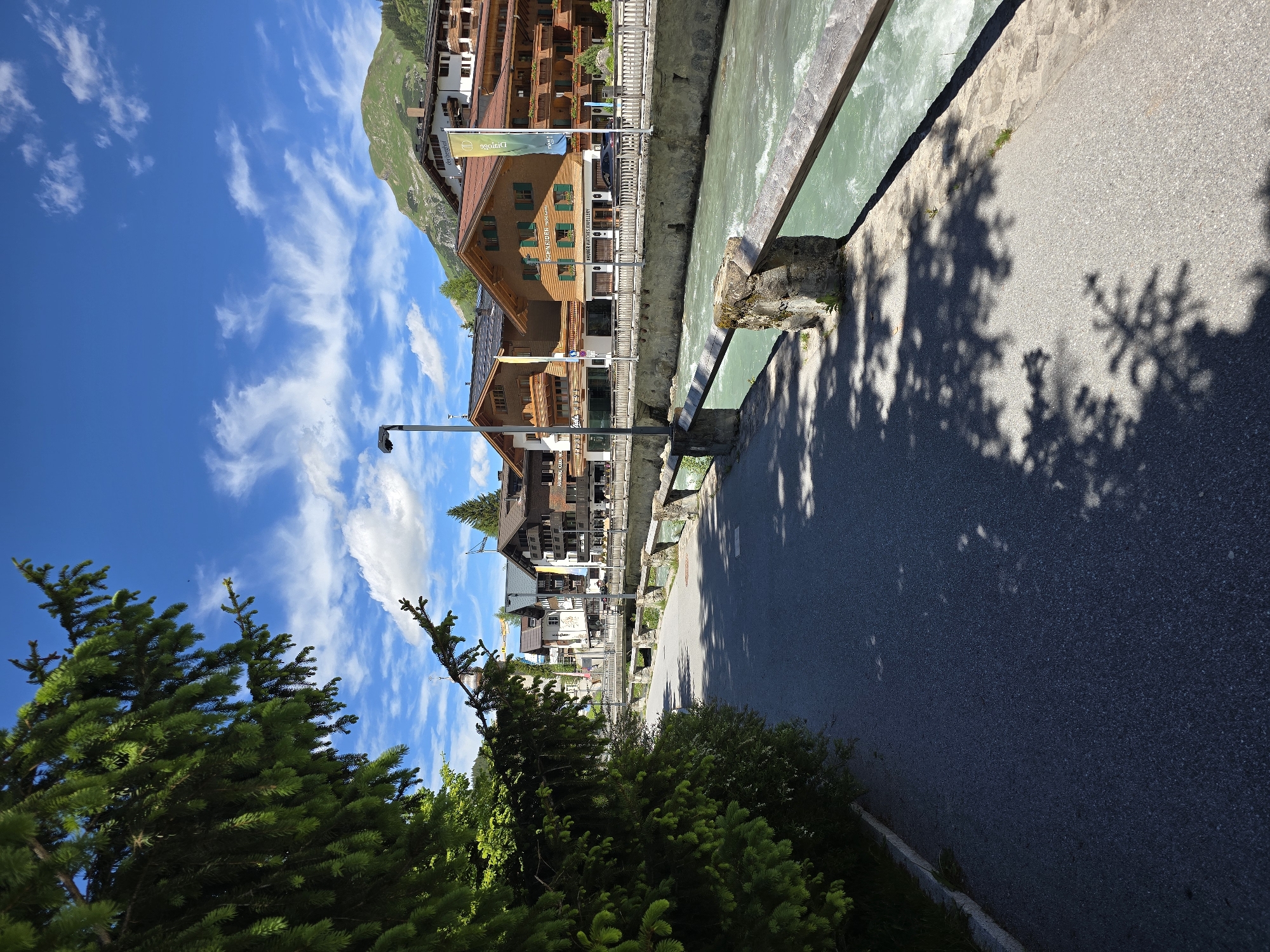 The image depicts a scenic view of the Lech river flowing through the village of Lech, Austria, on a bright, sunny day. On the left side of the frame, a lush green fir tree partially obscures the view, with its branches casting shadows on a paved walkway. The walkway runs alongside the river, which has a vibrant turquoise hue and a moderate current. A stone wall with a wooden railing borders the river.
Across the river, a row of traditional Austrian buildings stands, characterized by their wooden facades, peaked red-tiled roofs, and green shutters. Several of these buildings appear to be hotels or shops, with some displaying signage such as "SCHNEIDER INTERIOR DESIGN" and "GOLDSCHMIEDE Jewellery". A bridge can be seen spanning the river in the background, connecting the two sides of the village. Beyond the buildings, rolling green mountains rise under a clear blue sky dotted with scattered white clouds. The overall scene suggests a tranquil summer day in a picturesque alpine village. There are no people visible in the image.