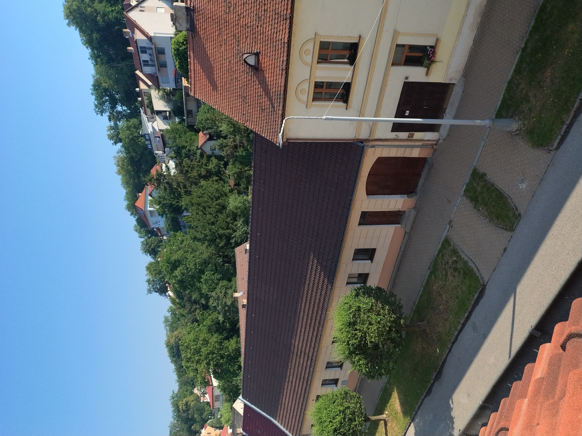 The image shows a view from a rooftop looking down onto a street and buildings in Hluboká nad Vltavou, Czechia. In the foreground, there are red roof tiles. The street below is paved with asphalt and has a sidewalk made of interlocking pavers, with patches of grass and small trees.
To the left of the street, there is a long, single-story building with a dark brown tiled roof. This building has several windows and a large double door. Two small, well-maintained green trees are planted in front of this building.
To the right of the street and slightly further back, there is a two-story building with a red tiled roof. This building is painted a light cream color with yellow trim. It features several windows, including two on the upper floor with arched detailing above them, and a small dormer window on the roof. A dark brown door is visible on the ground floor, and a window box with flowers is placed beneath one of the upper windows. A lamppost stands on the sidewalk in front of this building.
Behind these buildings, a dense green forest covers a hillside. Further up the hill, various houses and buildings with red and white roofs can be seen nestled among the trees. The sky is clear and blue, indicating a sunny day. No people are visible in the image.