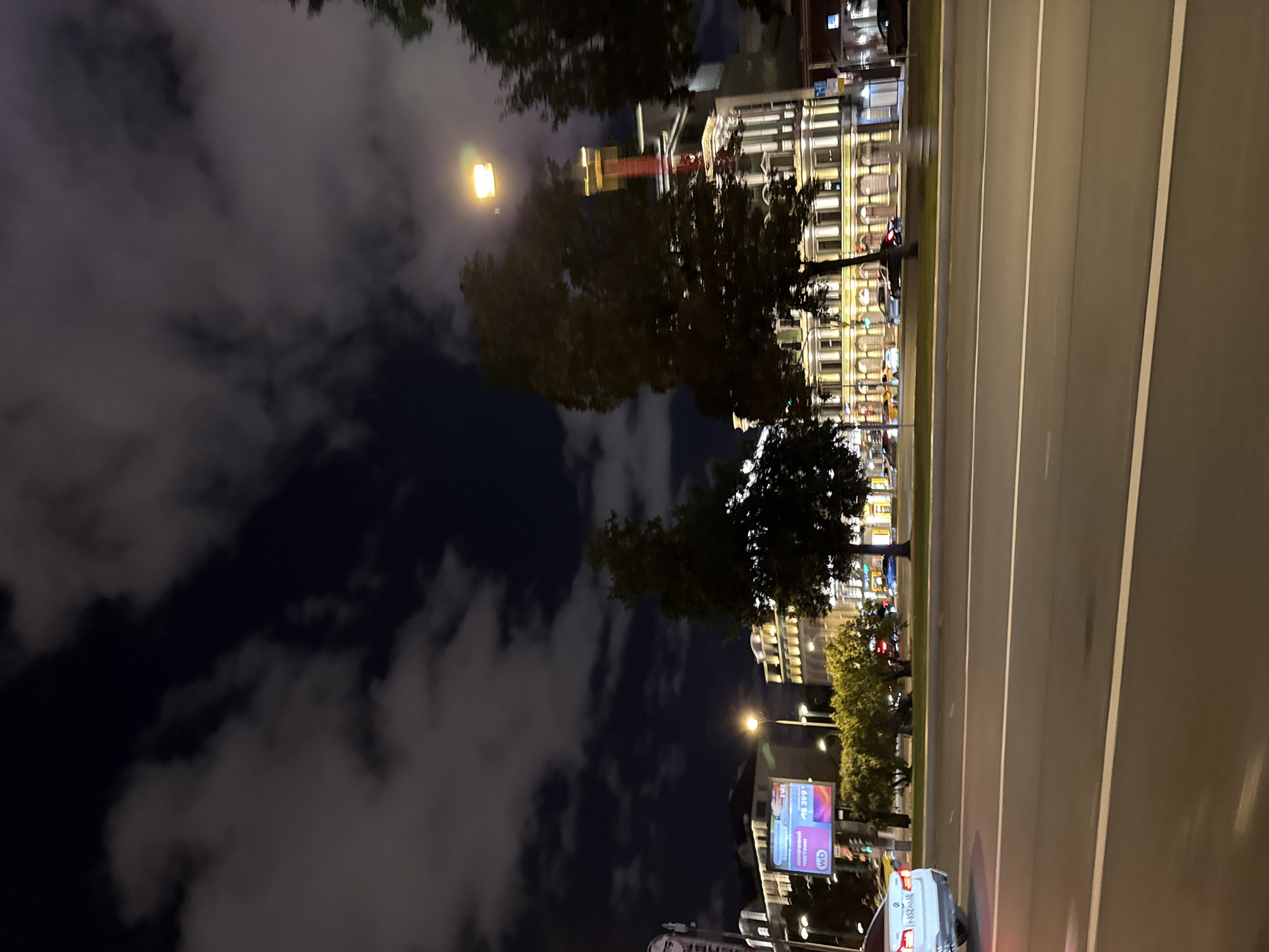 The image captures a nighttime street view in Moscow, Russia, likely along a major avenue. In the foreground, a multi-lane road is visible with white lane markings, indicating vehicular traffic. A white car is partially visible on the left side of the frame, appearing to be in motion.
Across the road, a row of illuminated buildings stands out. These structures exhibit classical architectural styles with arched windows and decorative facades, lit from within and by streetlights. One building on the right is taller and features a prominent, bright yellow light source at its upper section, possibly a beacon or a tall lamp. A large digital billboard displaying advertisements is situated between two buildings on the left.
Mature trees with dark foliage line the sidewalk in front of the buildings, with their branches illuminated by the artificial lights. The sky above is dark, with scattered, fluffy clouds tinged with gray and some areas of lighter illumination, suggesting the presence of moonlight or distant city lights filtering through. The overall atmosphere is that of a bustling urban environment at night, with the illuminated buildings and streetlights contrasting with the dark sky.
