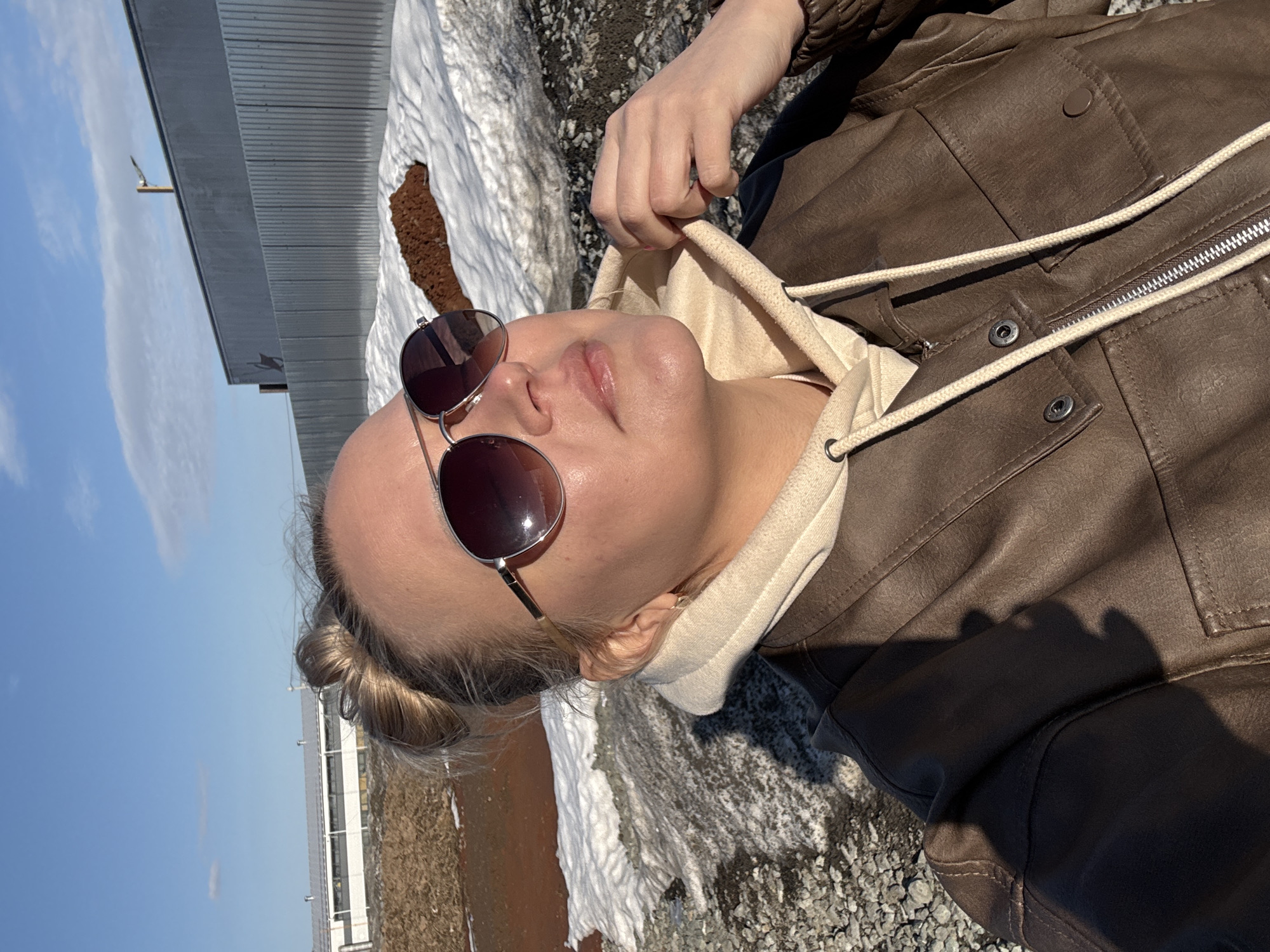 A woman wearing sunglasses, a tan hooded sweatshirt, and a brown leather jacket lies on the ground. The ground is mixed with gravel and patches of snow. In the background, a grey building is visible against a blue sky. Based on visual cues, the location is likely Izhevsk, Russia, due to the building design and remaining snow.