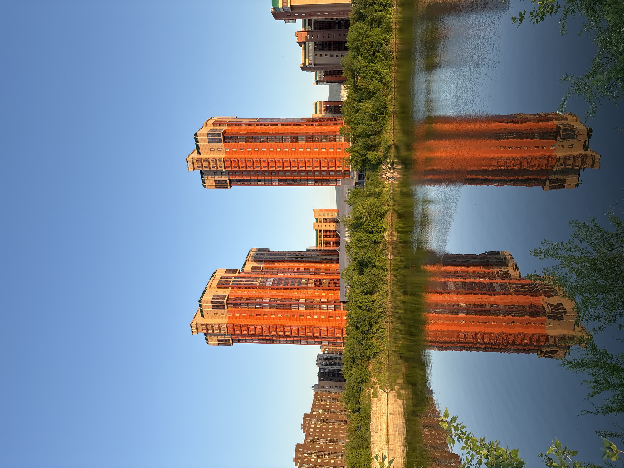 A static, landscape-oriented image captures a serene urban waterfront scene, viewed as if rotated 90 degrees clockwise (the actual content appears vertically aligned, with the sky on the left). When viewed correctly, tall, modern residential buildings with prominent orange-red and grey facades rise along the left bank of a calm body of water. Numerous windows are visible on the buildings. A line of lush green trees forms a natural barrier between the buildings and the water's edge.
The sun, likely in the late afternoon or early morning, illuminates the buildings with a warm, golden glow, making their colors pop against the clear, light blue sky. This warm light, along with the deep blue of the sky, is vividly reflected in the tranquil water, creating a near-perfect mirror image of the cityscape. The reflections are slightly softened and elongated by subtle ripples on the water's surface. In the background, beyond the prominent structures, other buildings of varying heights and colors can be seen stretching along the waterfront. The weather appears clear and sunny. The scene offers a blend of urban architecture and natural elements, characteristic of a developed city like Krasnoyarsk, Russia, situated by a river or lake. No people or visible text are present.
