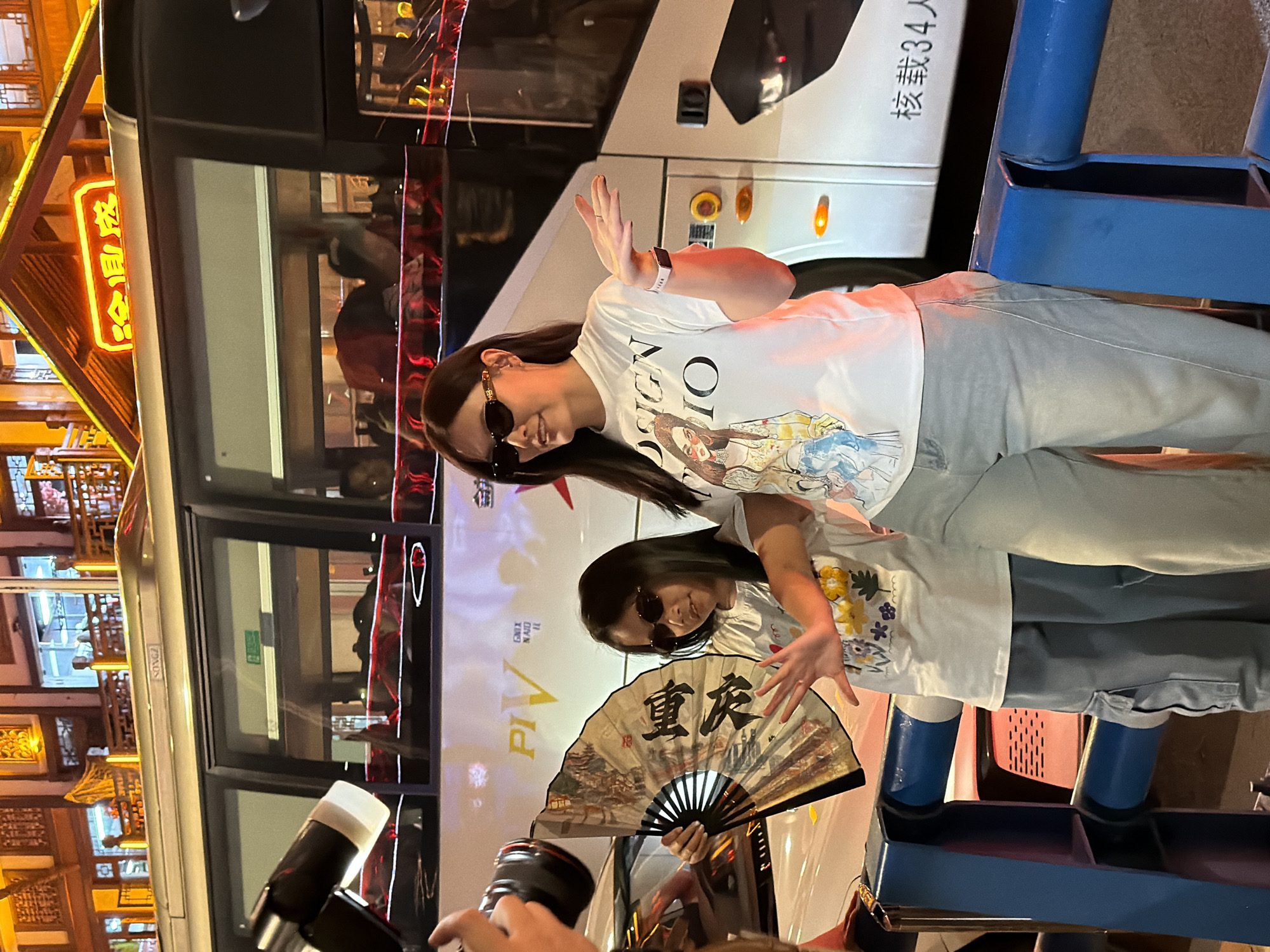 This is a nighttime photograph featuring two females posing for a picture, with a bus and illuminated buildings in the background. The image is taken outdoors, likely in a city environment.
The main subjects are two females, one taller and one shorter (possibly an adult and a child or two adults), both wearing white t-shirts, light-colored jeans, and sunglasses. The taller individual has long dark hair and is wearing a watch on her left wrist. Her white t-shirt has a partial visible design, including the letters "SIGN" and "IO". She holds her right hand up in a playful gesture. The shorter individual also has dark hair and holds a traditional Chinese fan with both hands, extending it towards the camera. The fan is decorated with a scenic painting and features prominent Chinese characters. Both subjects appear to be smiling beneath their sunglasses, suggesting a cheerful or confident mood. In the foreground, a person's hands holding a camera are visible, indicating they are taking the photograph.
Behind the subjects, a large white bus is parked, featuring Chinese characters "核载34人" (Rated capacity 34 people) and partially visible English text "SONGZ". Further in the background, to the left, are illuminated traditional Chinese-style buildings with glowing signs. One sign clearly displays the Chinese characters "冷圆成" (Lěng Yuánchéng), and other partial English text like "PIV", "GNIX", and "NAJO" is visible on a backdrop behind the shorter person. Blue metal structures, possibly public seating or barriers, are visible on the right side of the image.
The time of day is clearly night, as evidenced by the dark surroundings and prominent artificial lighting from the building signs and vehicle lights. The Chinese characters on the bus, building signs, and specifically "重庆" (Chóngqìng) on the fan, strongly indicate the location is in China, consistent with the provided context of Yuzhong District.