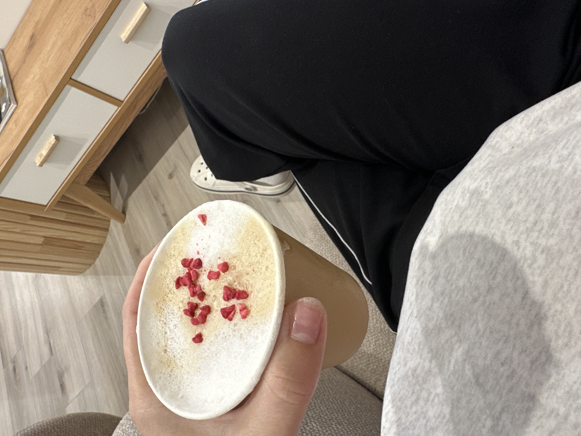 A person wearing black pants and white sneakers is holding a beige paper cup with a white foamy drink topped with red pieces. The location appears to be indoors, near a light-colored wooden cabinet or drawer unit. The image does not provide sufficient information to definitively identify the location as Perm, Russia.