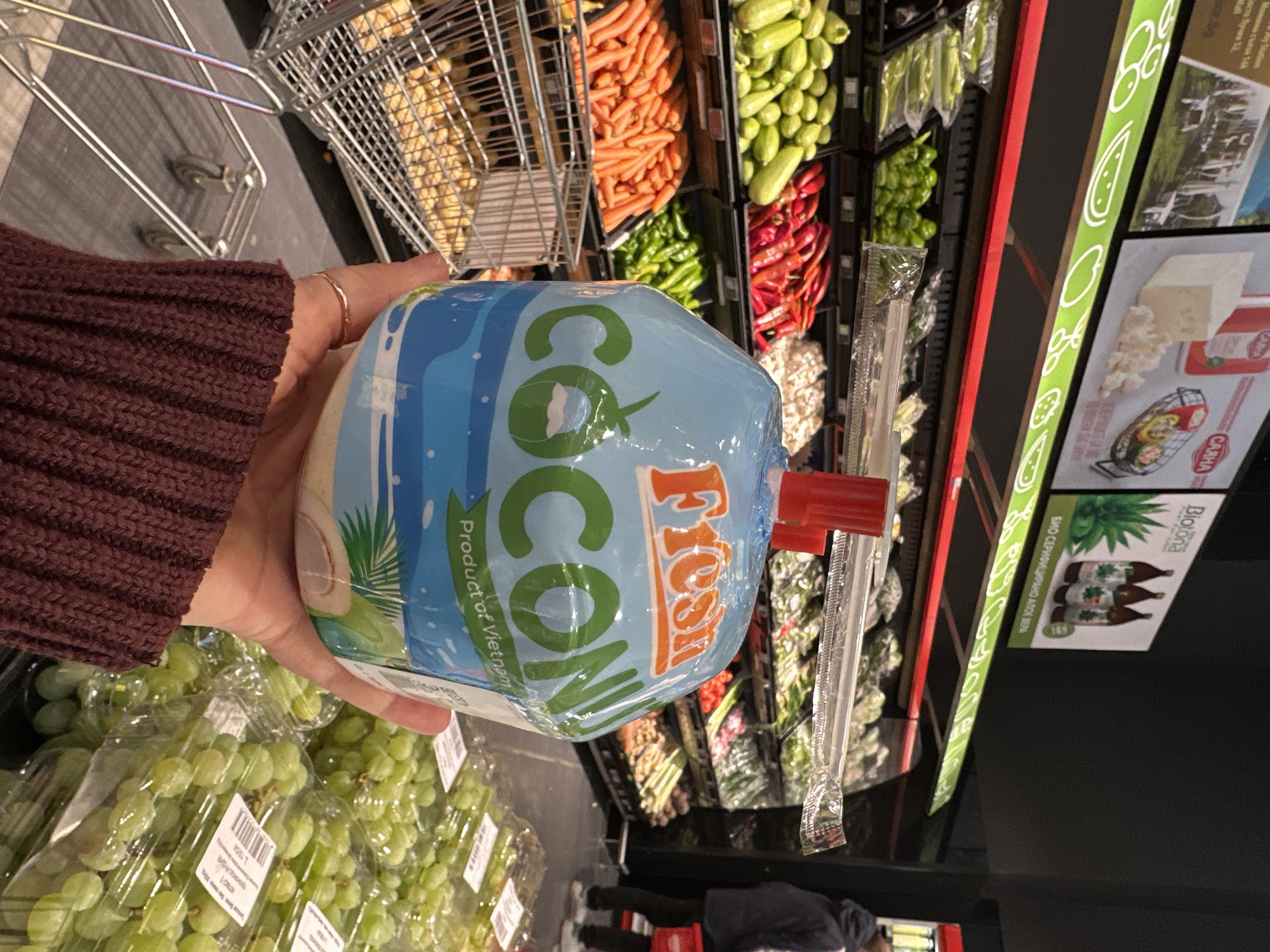 A person in a maroon sweater holds a Fresh Coconut drink in a grocery store. The packaging indicates it is a product of Vietnam. A packaged straw is taped to the side. A shopping cart, filled with potatoes and carrots, sits in the background, along with other produce. The image includes a store shelf displaying cheese and aloe vera juice products, with Cyrillic text possibly indicating a location in Sofia, Bulgaria. Grapes sit at the bottom of the image.