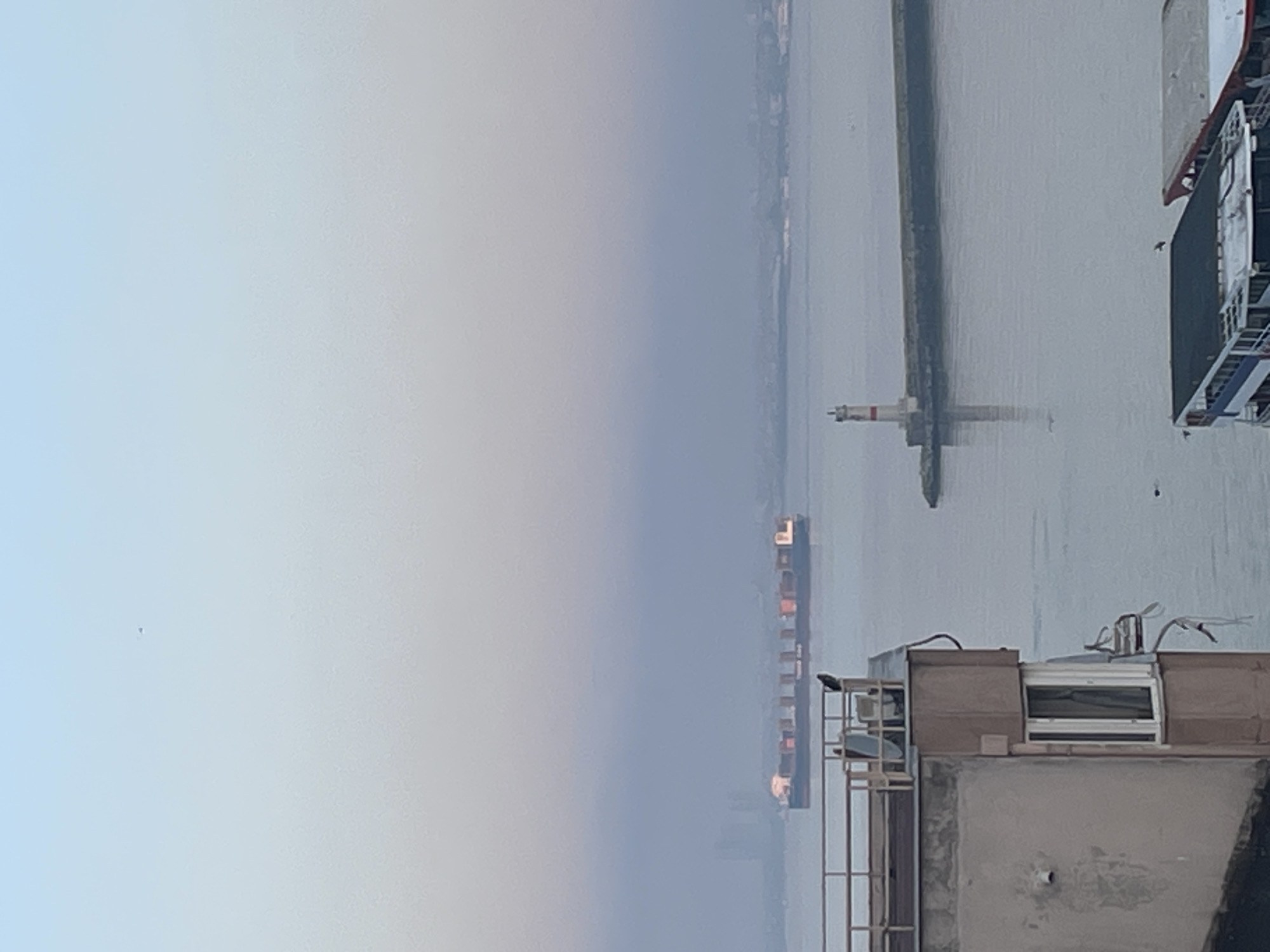 The image depicts a hazy view of the Bosphorus strait in Kadıköy, Turkey, during what appears to be early morning or late evening due to the soft, diffused light. In the foreground, a section of a building is visible on the left, with a balcony railing and a satellite dish. A bird is perched on the railing. The strait is calm, with a large container ship sailing in the middle ground. To the right, a lighthouse stands on a concrete breakwater. In the far background, the silhouette of a city skyline is faintly visible through the mist. Several boats are docked in the foreground at the bottom right of the image. The overall atmosphere is serene and quiet.