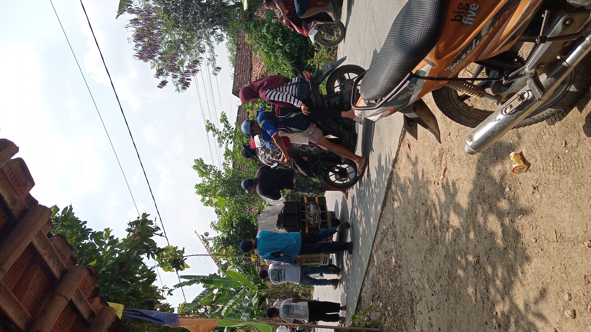 The image depicts a street scene in Belor, Indonesia, on a sunny day. Several individuals are present, some on foot and others on motorcycles. In the foreground, a partially visible orange and black motorcycle is parked. The background reveals a residential area with houses and lush vegetation, characteristic of a rural or suburban Indonesian setting.
A group of men are gathered around a small, motorized cart loaded with white objects, possibly goods or equipment. One man in a blue shirt is standing next to the cart, and another in a black shirt is behind it. A third man, wearing a patterned shirt, stands beside them, holding what appears to be a small Indonesian flag.
A motorcycle is positioned in the center of the frame, with two people riding it. The male driver wears a blue and black shirt and shorts, and a woman with a red headscarf and a striped shirt is the passenger. Another motorcycle, partially visible on the right, is ridden by two individuals, one wearing a red shirt.
Several other people are seen on the left side of the image, including a young boy in a white t-shirt and black pants, and two men in white t-shirts. The street is paved, and the ground on the left is a dusty, unpaved surface. Overhead, power lines are visible against a partly cloudy sky. The overall impression is one of everyday activity in a local community.