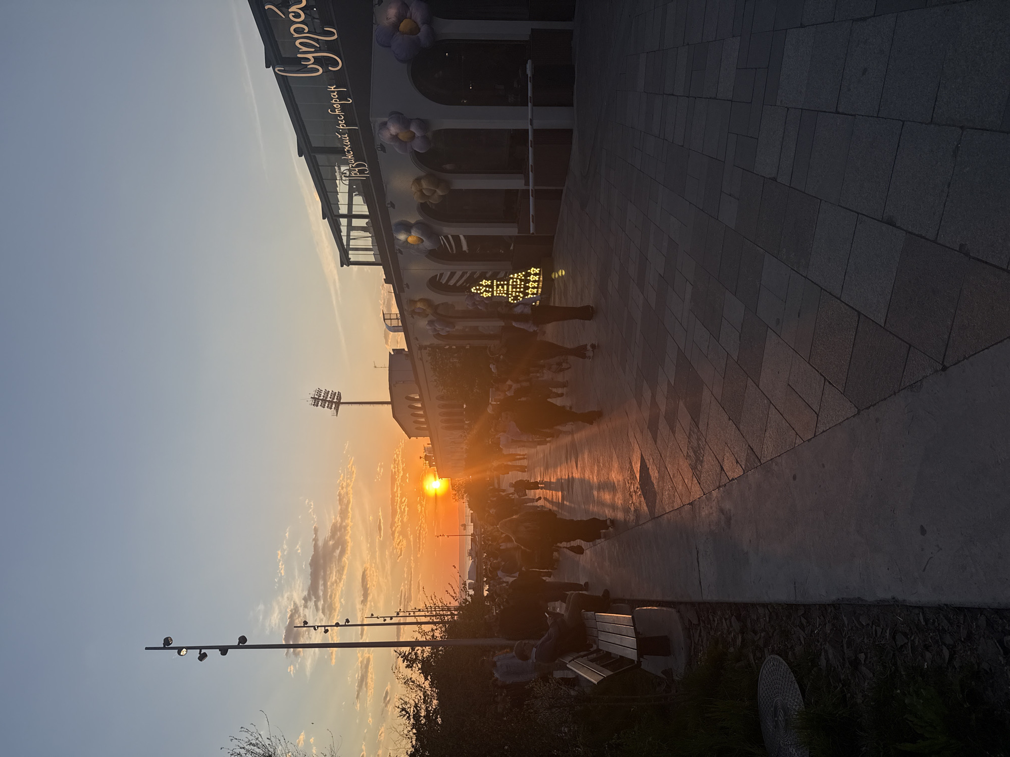 The image captures a sunset scene in Vladivostok, Russia. The sun is setting over the water, casting a warm orange glow across the sky and reflecting off the paved walkway. A modern building, possibly a restaurant, is visible on the right side of the image, adorned with floral decorations and signage in a Cyrillic alphabet. People are walking along the walkway, their silhouettes blurred by the sunlight. In the foreground, there is a bench with people seated, enjoying the sunset. Streetlights line the edge of the walkway, contrasting with the natural light. In the background there is a stadium.