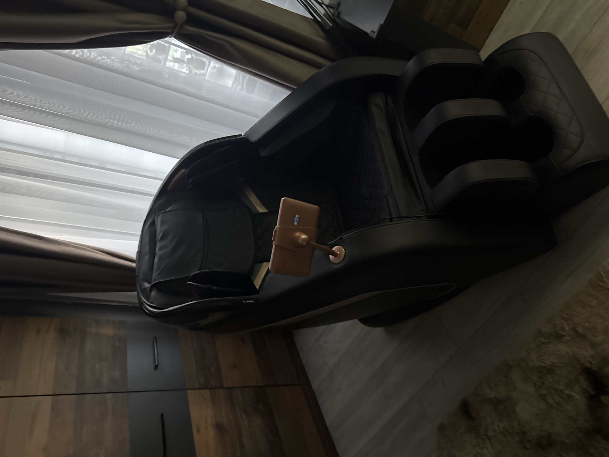 The image shows a black massage chair in an indoor setting. The chair is positioned near a window with light filtering through white horizontal blinds. The curtains are brown. To the left of the chair is a piece of furniture with dark wood and black accents. The floor is light-colored and appears to be wood or laminate. A tan rug is located to the right of the chair. A tablet is visible on the side of the chair. The location is likely inside a building, possibly a home or office in Kostinbrod, Bulgaria. There is no indication of any people.