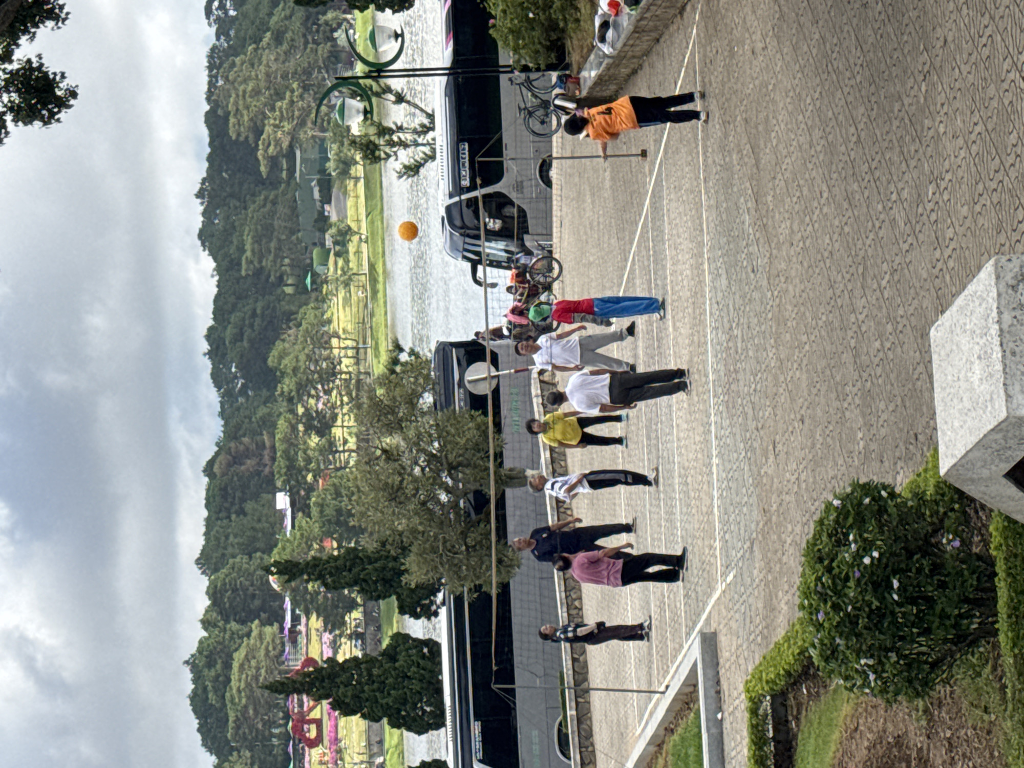 This image depicts an outdoor recreational scene in Da Lat, Vietnam, set in a paved public area adjacent to a lake, likely Xuan Huong Lake. The sky is overcast. A group of approximately ten adults, appearing to be middle-aged to elderly, are playing volleyball on a rudimentary court defined by white lines on the patterned gray pavement. An orange volleyball is suspended in mid-air above a makeshift net. One player in an orange shirt and black pants is positioned on the right, holding a paddle-like object, having just struck the ball. Other players, dressed in casual sportswear, are positioned on both sides of the net.
In the mid-ground, two large silver and black tour buses are parked parallel to the lake, facing away from the viewer. Several bicycles are parked or being ridden near the buses. The lake surface is visible behind the buses and net. Beyond the lake, lush green hills covered with dense trees extend into the distance. On the far left, across the lake, colorful floral displays indicative of a flower garden are visible. Two prominent green lampposts with twin white globes stand on the right side of the frame. The foreground features concrete ledges, manicured hedges, and a small area of grass.